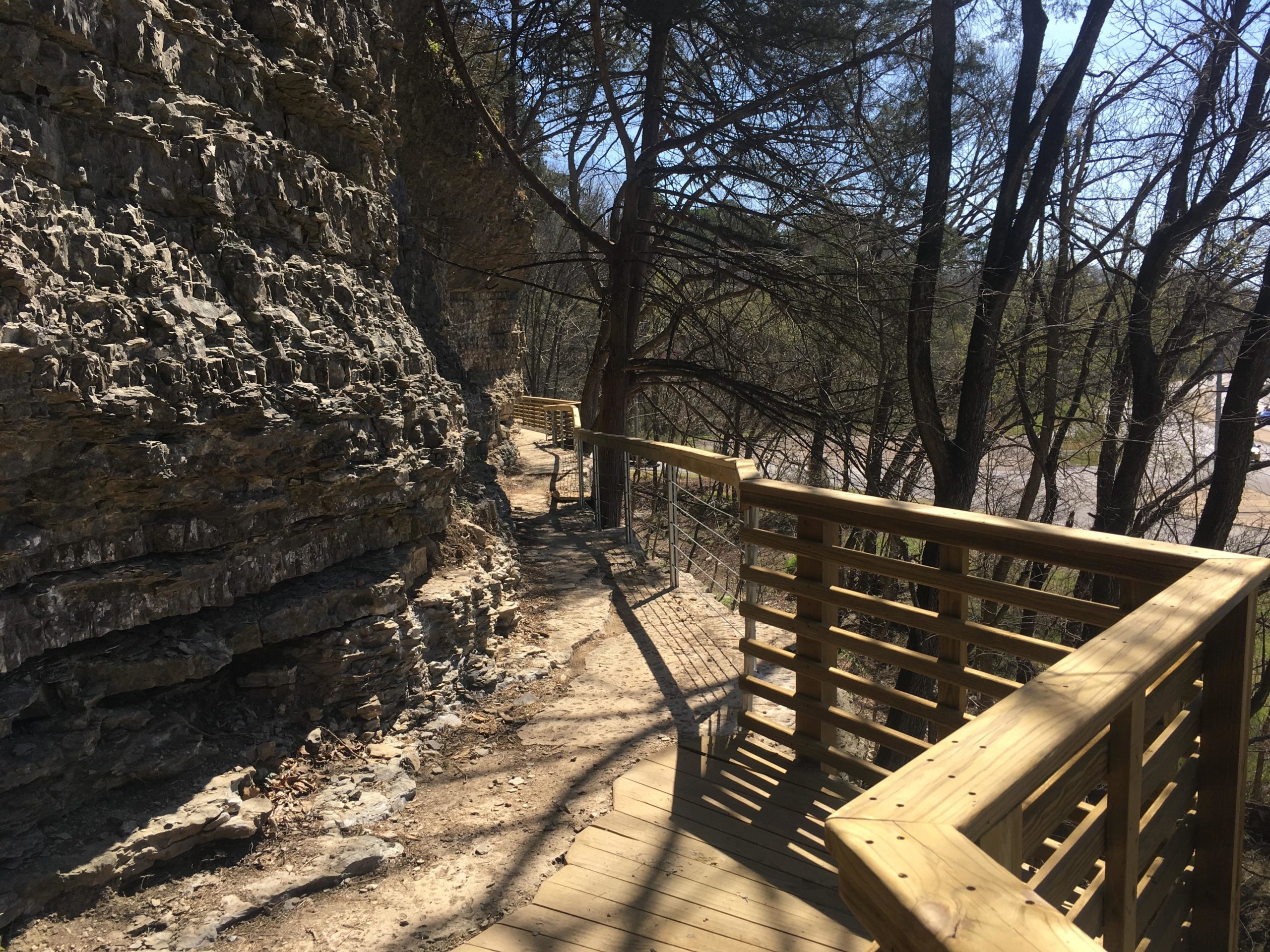 A wooden walkway meanders alongside a rocky cliff, surrounded by trees. The sun casts shadows on the path, creating a serene natural setting. In the background, a road is visible, adding a sense of access to the outdoor space. Back 40 mountain bike trail.