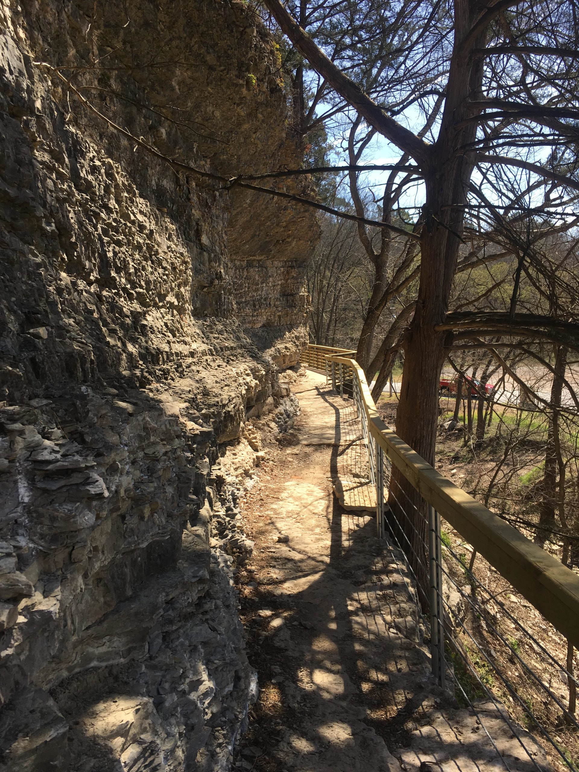 A narrow pathway alongside a rocky cliff, bordered by a wooden railing. Sunlight filters through trees, casting shadows on the ground. Back 40 mountain bike trail.