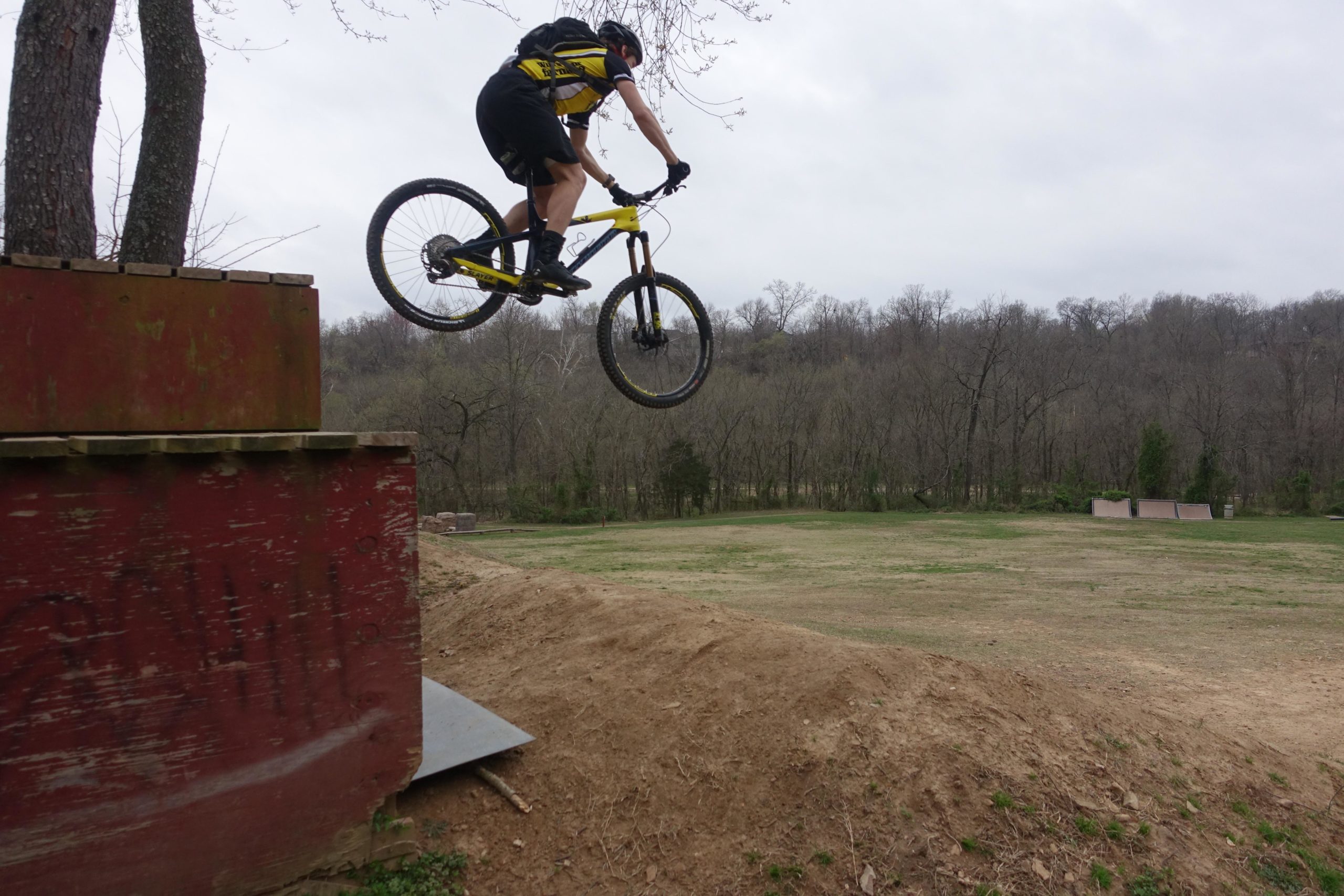 A mountain biker in a yellow and black outfit jumping off a wooden ramp, airborne over a dirt hill, with a cloudy sky and wooded area in the background. Slaughter Pen Trail mountain bike trail.