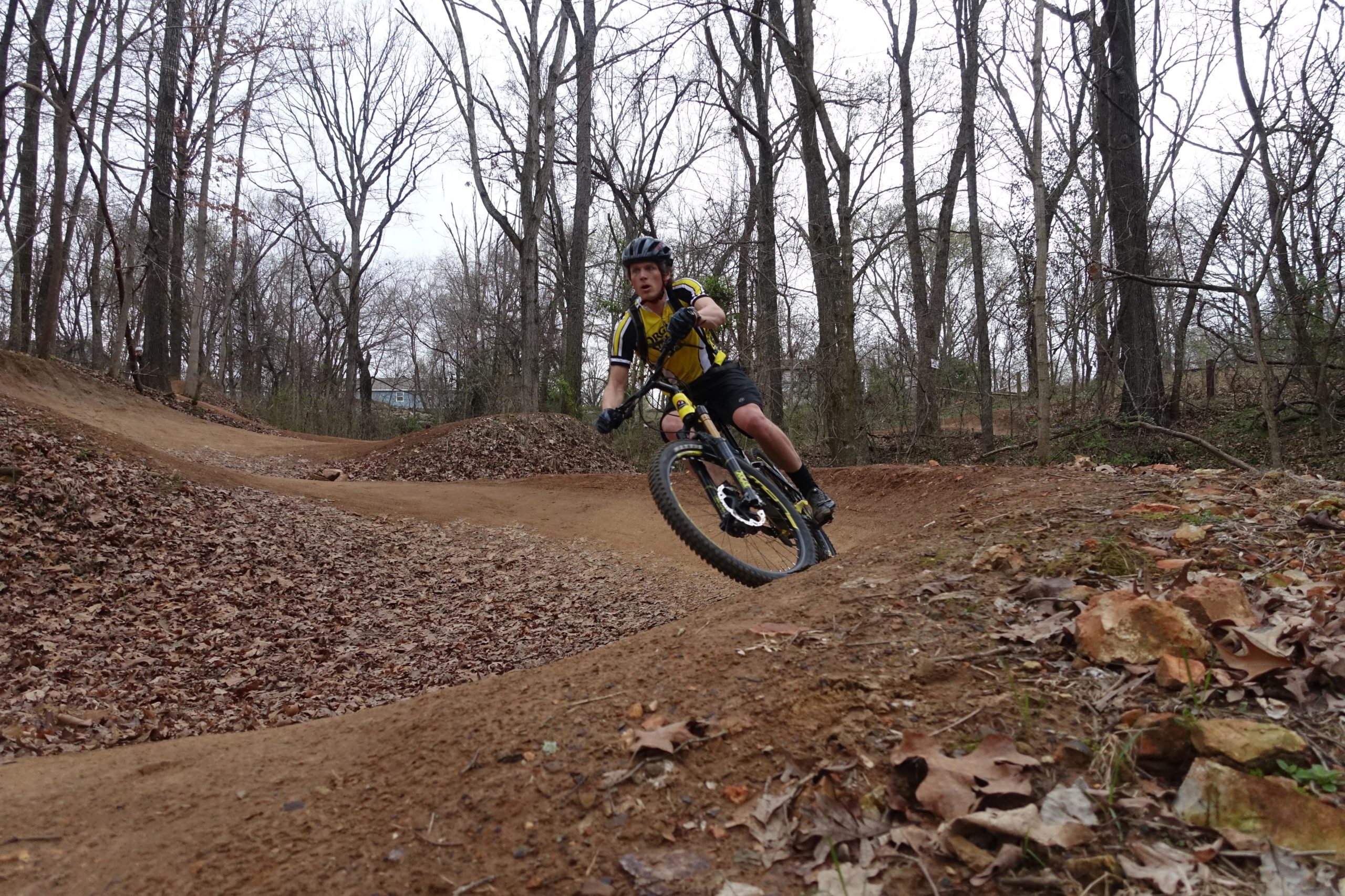 A mountain biker in a yellow jersey and black shorts leans into a curve on a dirt trail surrounded by leafless trees. The ground is covered with leaves, and there are dirt mounds alongside the path, indicative of a biking trail designed for jumps and turns. The scene is set in a wooded area on an overcast day. Slaughter Pen Trail mountain bike trail.