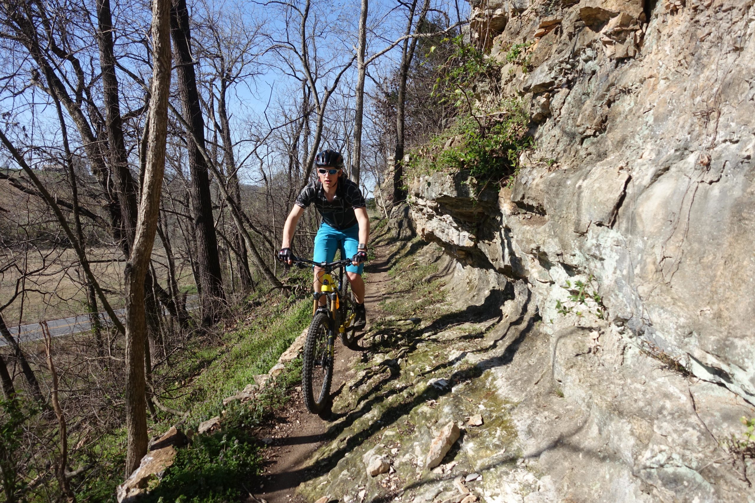 A person riding a mountain bike on a narrow dirt trail alongside a rocky cliff, surrounded by bare trees and a clear blue sky. The rider is wearing a helmet and sunglasses, dressed in a black shirt and blue shorts. Back 40 mountain bike trail.