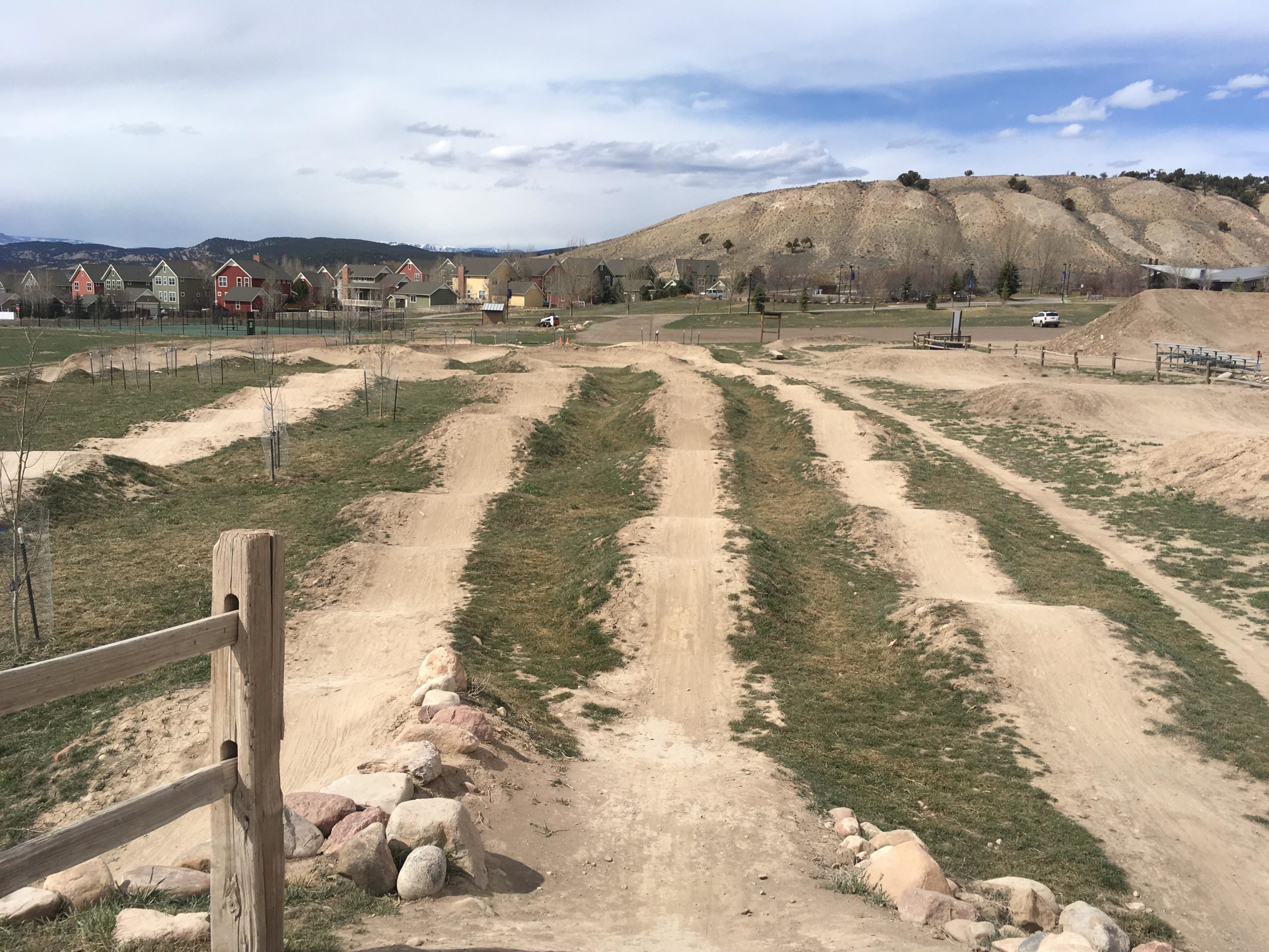 A dirt bike park features several dirt mounds and trails, with grassy areas between them. In the background, residential homes can be seen, set against a backdrop of rolling hills under a partly cloudy sky. A wooden fence and a rocky border are visible in the foreground. Eagle Bike Park mountain bike trail.