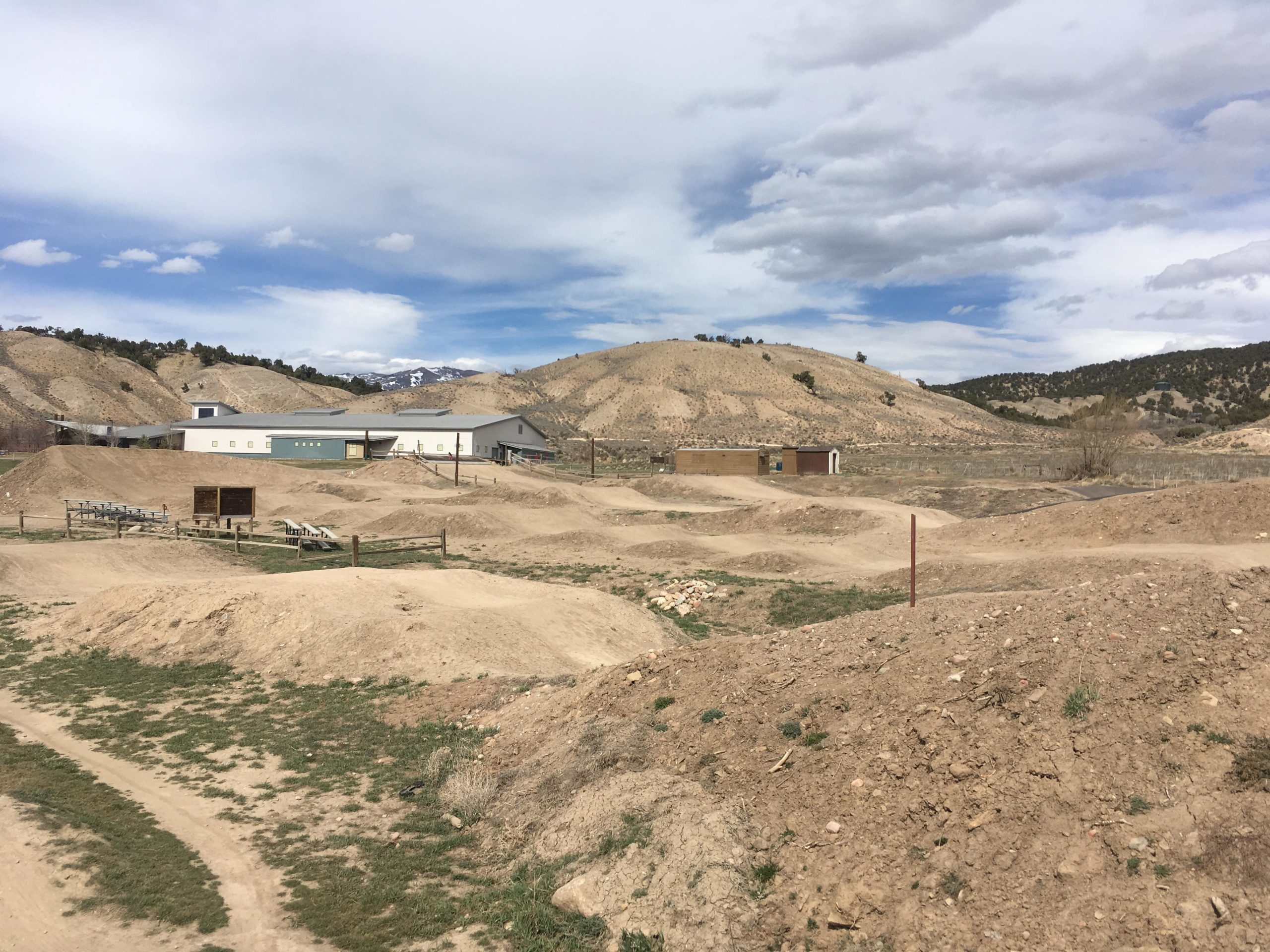 A landscape featuring dirt mounds and slopes, with a few small buildings in the background. The area appears to be a rural or outdoor setting, with rolling hills and some greenery visible among the dirt. The sky is partly cloudy, and there are snow-capped mountains in the distance. The foreground includes a dirt path winding through the area. Haymaker Trail mountain bike trail.