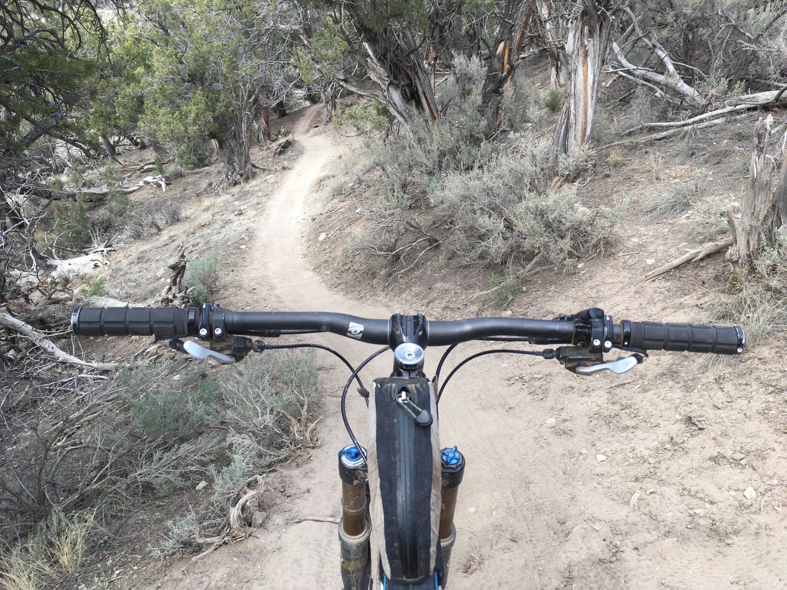 Close-up view of mountain bike handlebars with a dirt trail winding through a natural landscape in the background, featuring sparse vegetation and rocky terrain. Haymaker Trail mountain bike trail.