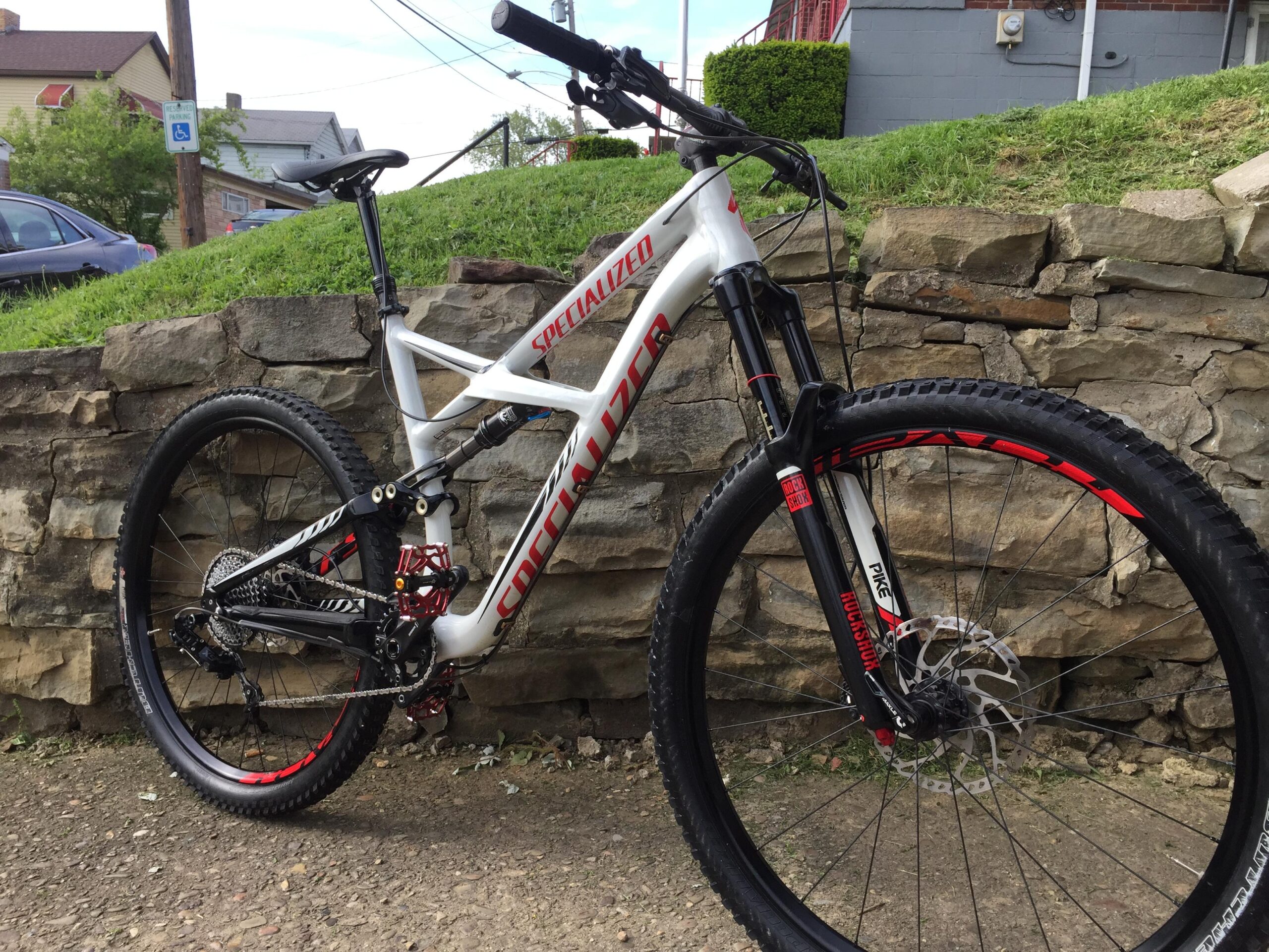 Specialized Enduro Comp: A white and red Specialized mountain bike leaning against a stone wall, featuring thick tires, a dual suspension system, and a visible disc brake. The background shows grass and a residential area.