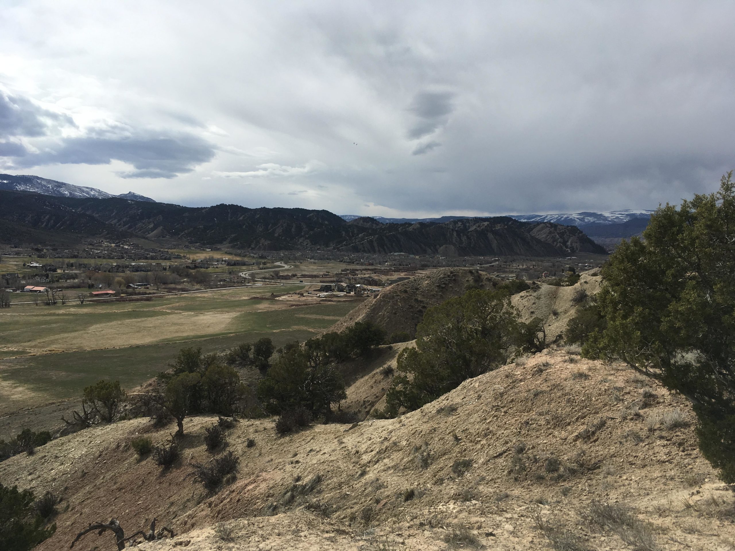 A scenic view of a valley surrounded by mountains, featuring rolling hills, patches of grassland, and scattered trees. The sky is overcast with gray clouds, and snow-capped peaks are visible in the distance. A winding road cuts through a small settlement in the valley. Haymaker Trail mountain bike trail.