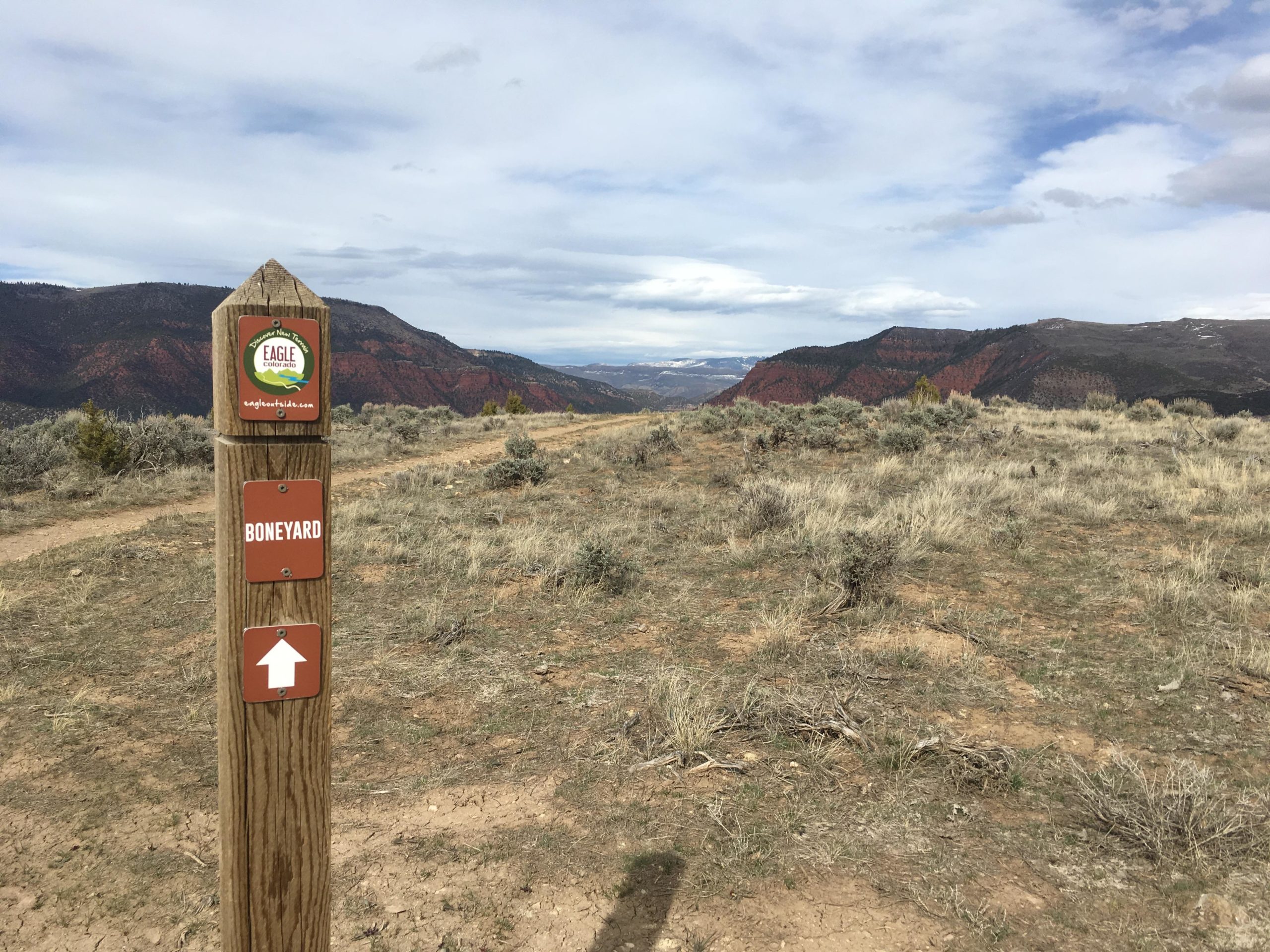 Wooden directional sign at a trailhead with "Eagle" and "Boneyard" markers, overlooking a scenic view of red rock formations and rolling hills under a cloudy sky. Sparse vegetation covers the ground. The Boneyard mountain bike trail.