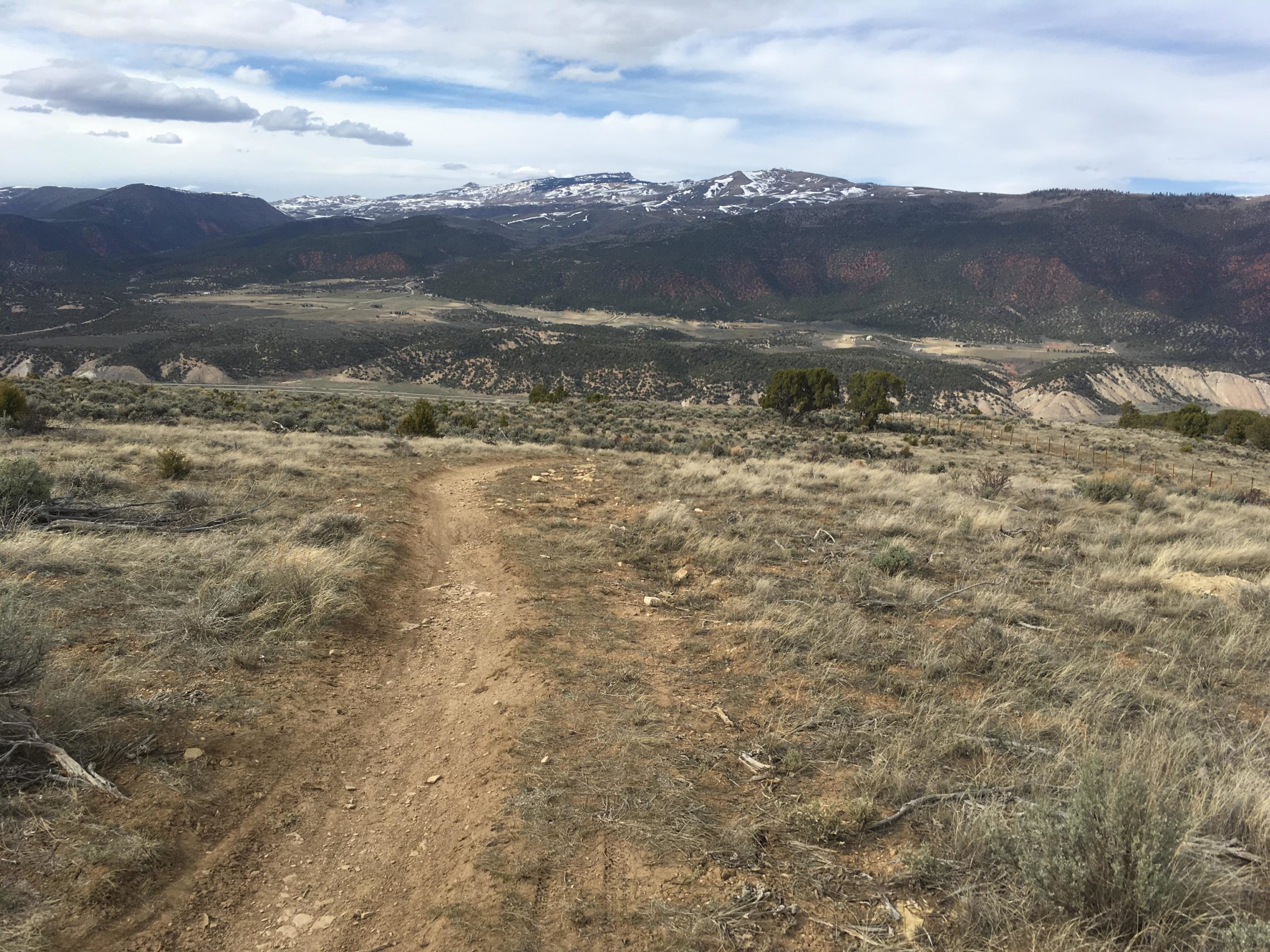A winding dirt trail through an open landscape, leading towards distant mountains covered in snow. The foreground features dry grasses and sparse vegetation, while the background showcases rolling hills and a valley beneath a partly cloudy sky. The Boneyard mountain bike trail.