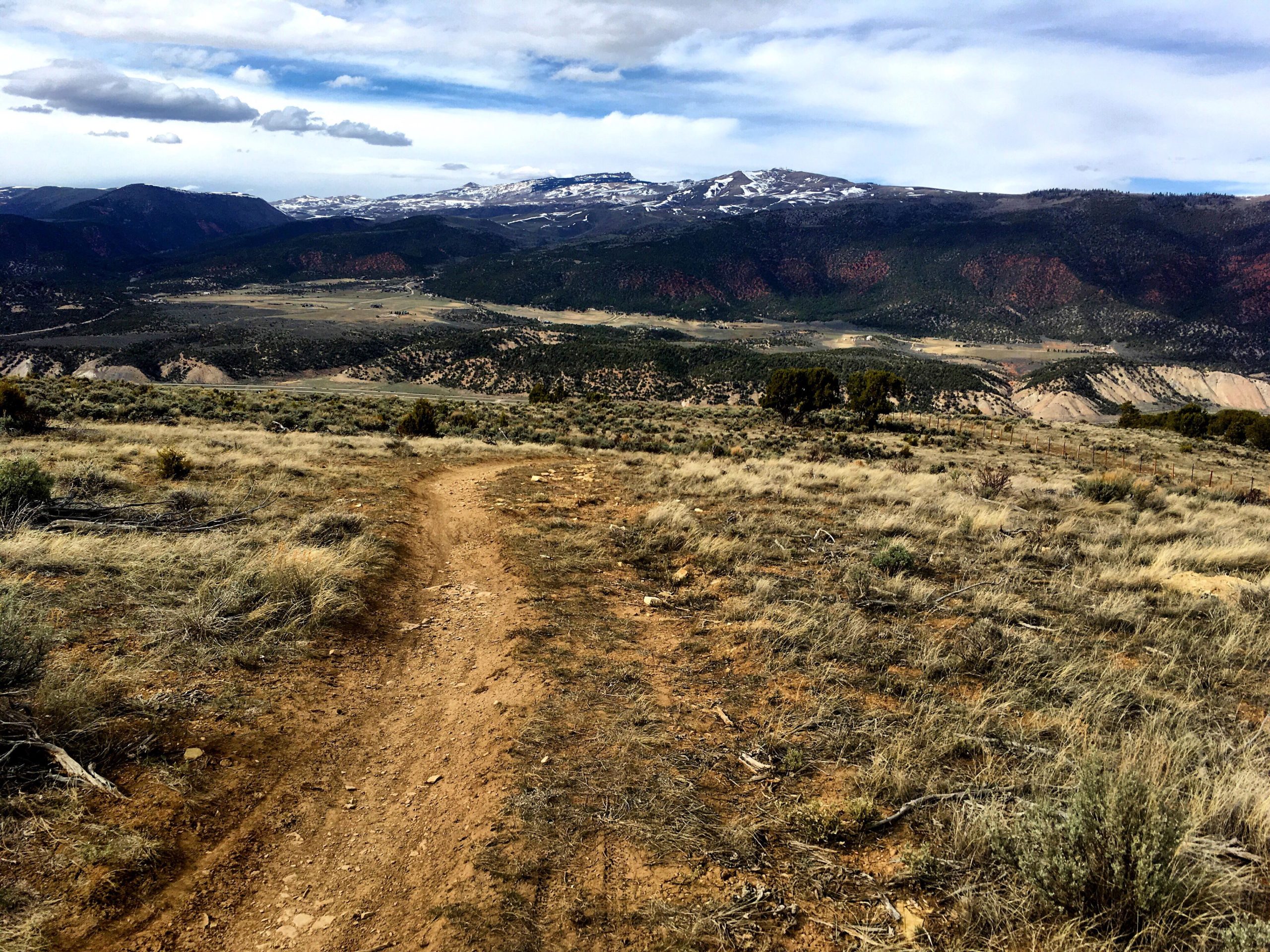A dirt path winding through grassy terrain, leading towards a picturesque landscape of rolling hills and snow-capped mountains under a cloudy sky. The foreground features sparse vegetation and dried twigs, while the distant hills display a mix of green and reddish tones. The Boneyard mountain bike trail.