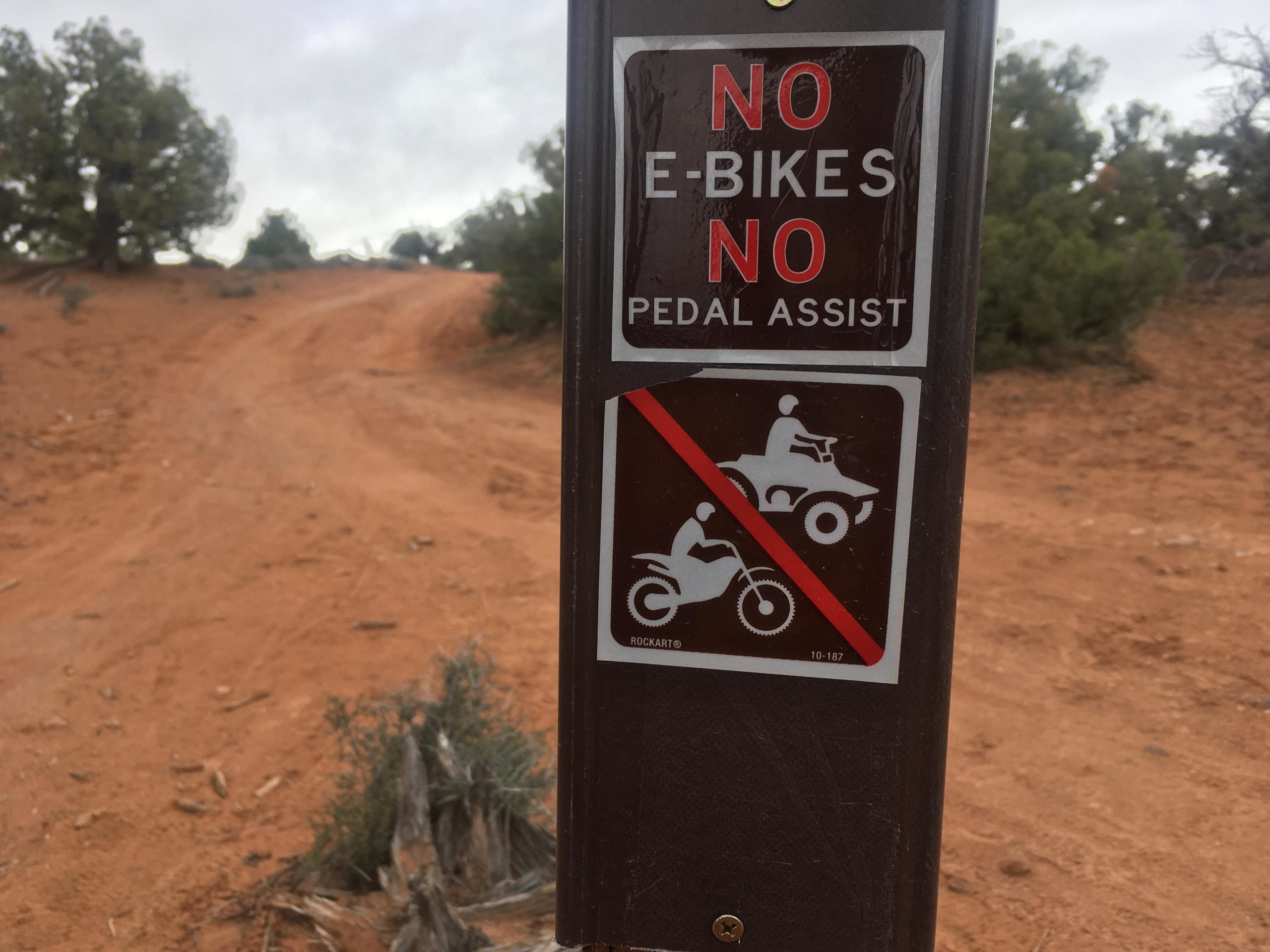 Sign prohibiting e-bikes, pedal assist bikes, and motorized vehicles (ATVs and dirt bikes) on a dirt road in a natural setting, with trees and a sandy path visible in the background. Navajo Rocks mountain bike trail.