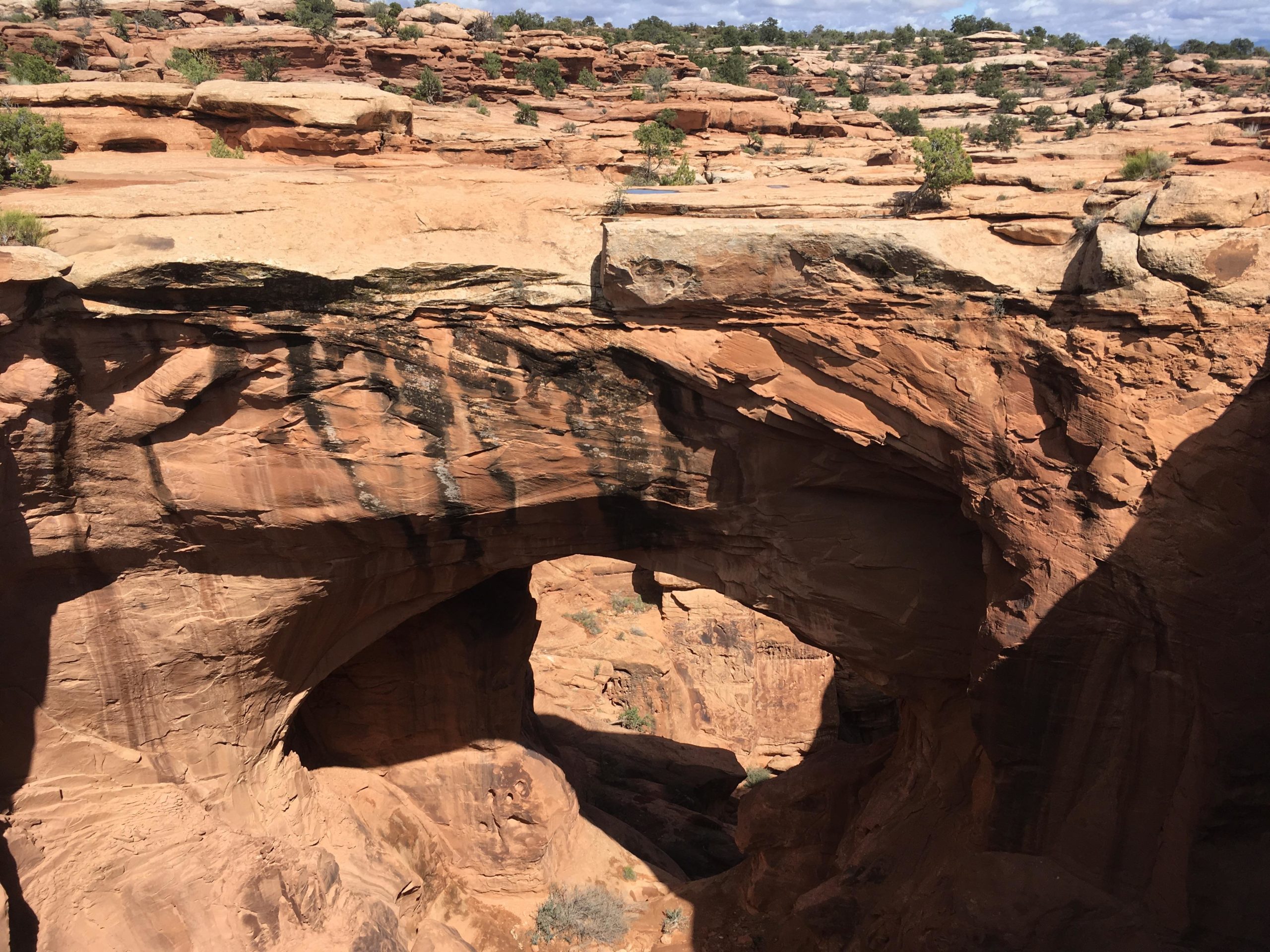 A natural stone arch formed by erosion in a desert landscape, surrounded by red rock formations and sparse vegetation under a blue sky with scattered clouds. Gemini Bridges mountain bike trail.
