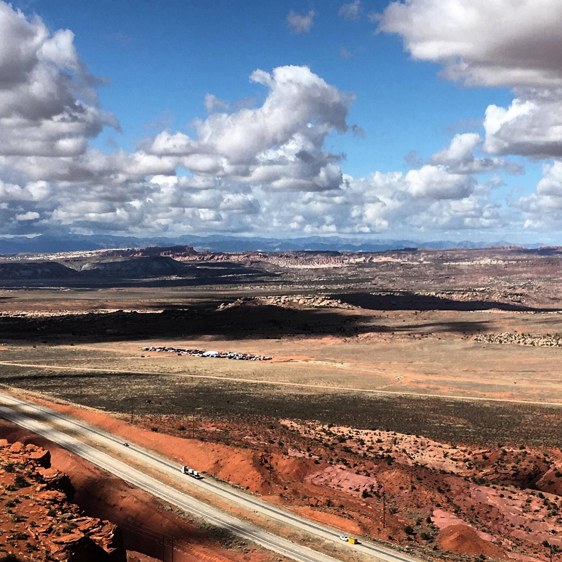 A wide view of a vast desert landscape featuring red and brown rocky terrain under a cloudy blue sky. A highway runs through the foreground, and a distant line of vehicles can be seen along the horizon. Rugged mountains are visible in the background. Gemini Bridges mountain bike trail.
