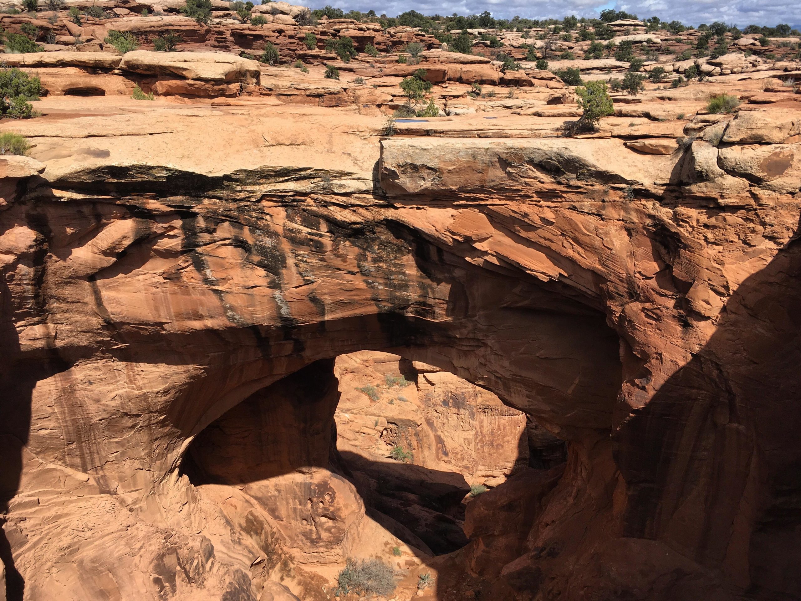 A scenic view of a rocky canyon landscape featuring a large natural archway formed from rust-colored sandstone. The image captures the contrast between the smooth surfaces of the arch and the rugged terrain, with scattered shrubs and sparse vegetation in the surrounding area. The sky above is partly cloudy, reflecting the natural beauty of the arid environment. Gemini Bridges mountain bike trail.