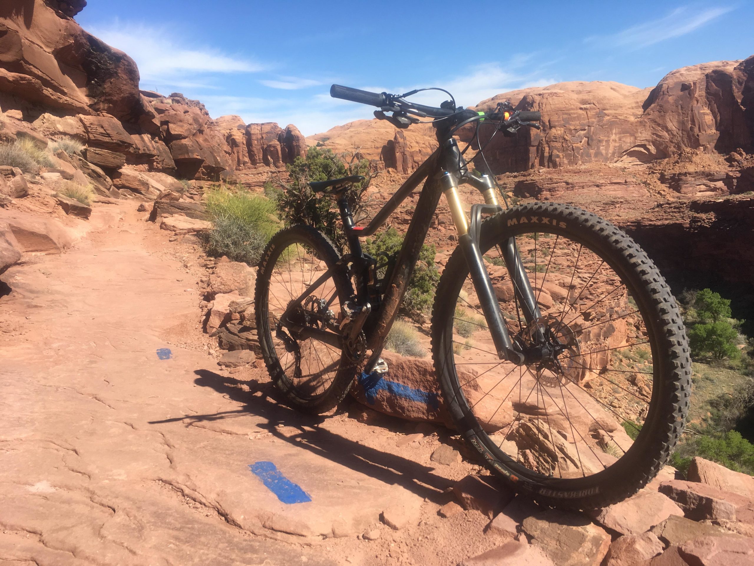 Mountain bike resting on a rocky trail surrounded by red rock formations and blue skies. The bike, equipped with rugged tires, is positioned next to blue trail markers on the ground, indicating the path ahead.  Captain Ahab mountain bike trail.