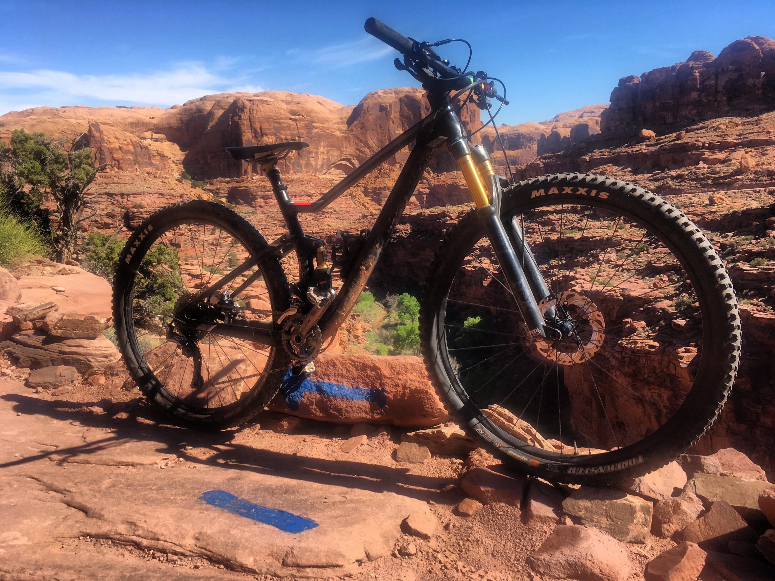 A mountain bike parked on a rocky trail with a scenic backdrop of red rock formations and a clear blue sky. The bike is poised at the edge of a canyon, highlighting its rugged tires and suspension system. Blue trail markings are visible on the ground, indicating a designated mountain biking path. Captain Ahab mountain bike trail.