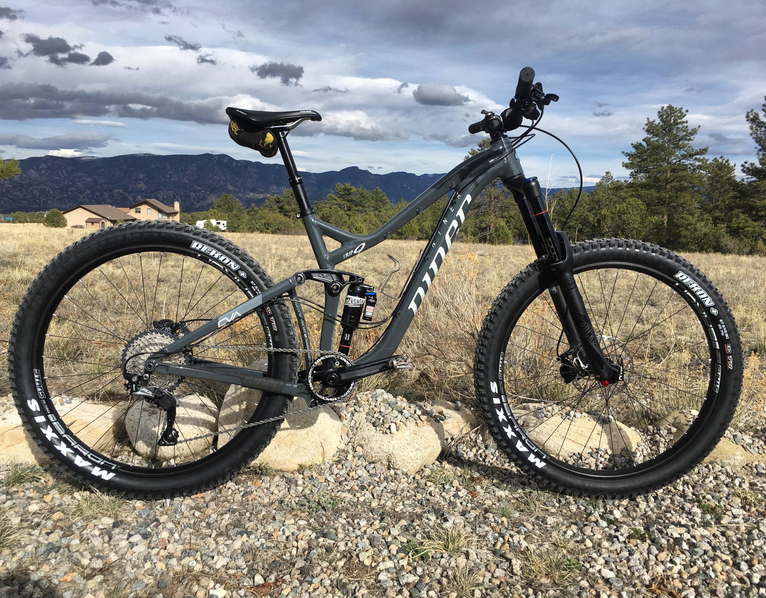 Niner RIP 9: A mountain bike with a gray frame, featuring thick tires, a front suspension fork, and a black seat. It is positioned on rocky terrain with a scenic background of mountains and cloudy skies. The bike is angled to showcase its components and is surrounded by grass and a few scattered rocks.