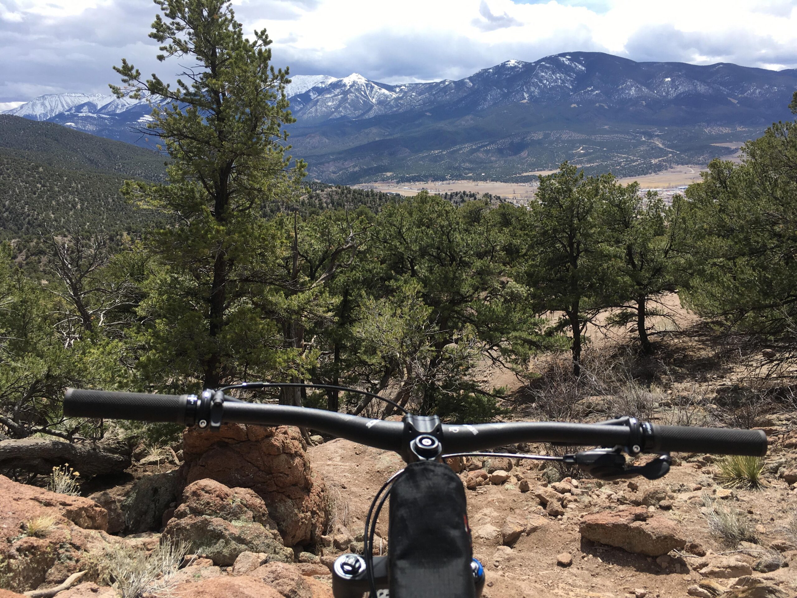 Niner RIP 9: A mountain biker's view from the handlebars, overlooking a mountainous landscape with green trees and rocky terrain. Snow-capped peaks are visible in the distance under a partly cloudy sky.