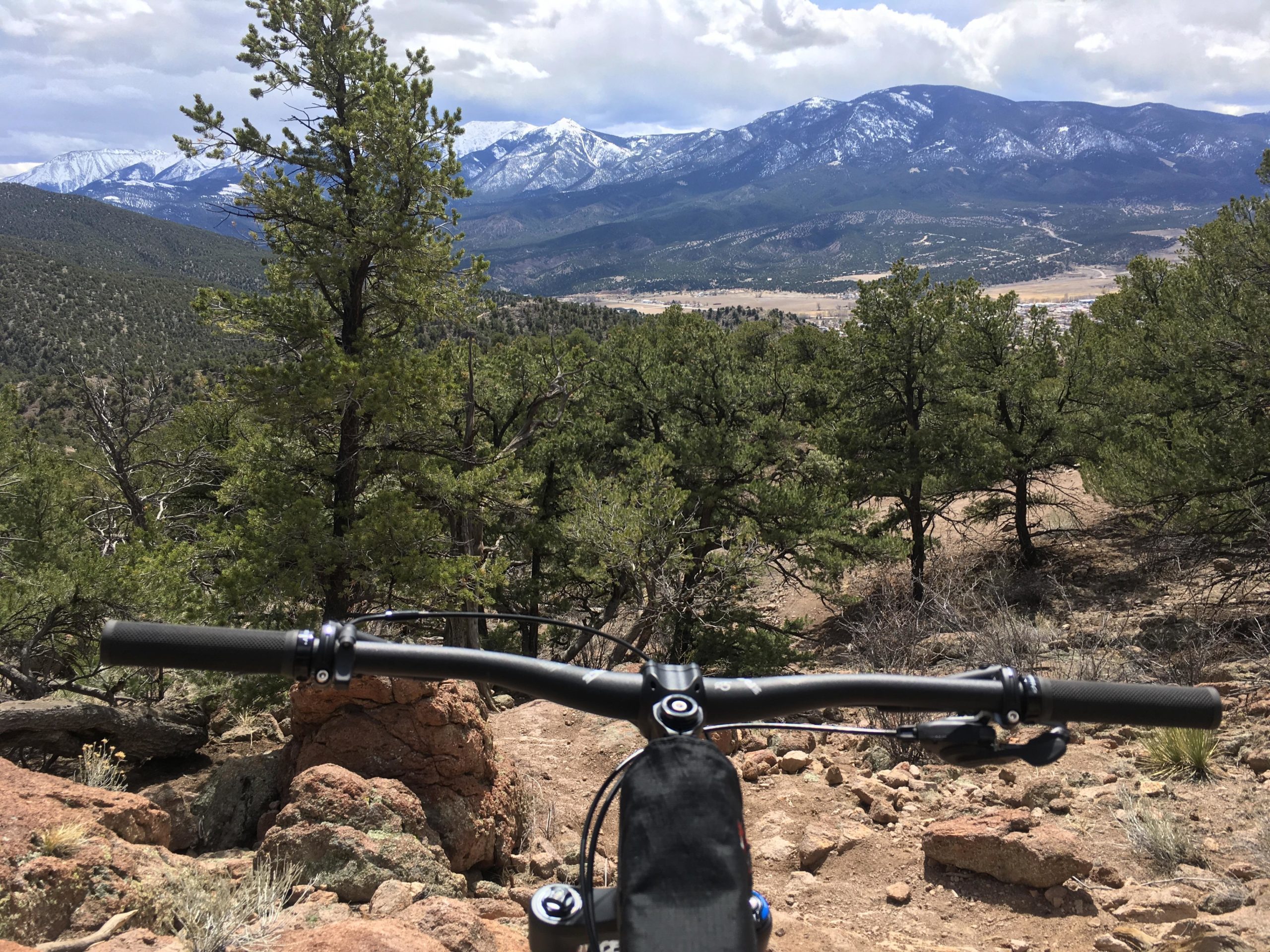 A mountain biker's view from the handlebars, overlooking a scenic valley surrounded by forested hills and snow-capped mountains under a partly cloudy sky. Unkle Nazty mountain bike trail.