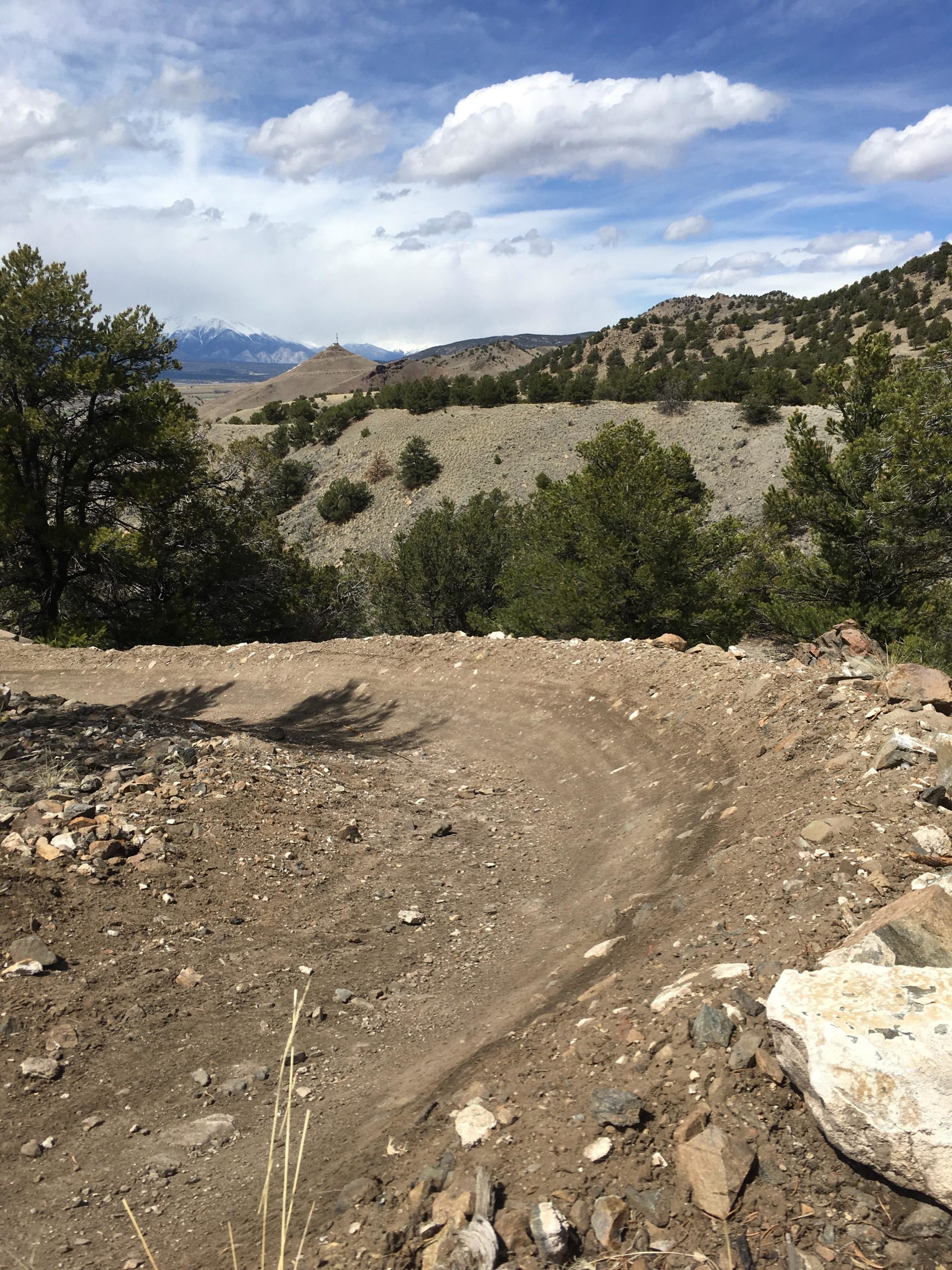 A winding dirt trail curves through a rocky landscape, surrounded by sparse vegetation and trees. In the distance, rolling hills and mountains are visible under a partly cloudy sky. The scene captures the natural beauty of a mountainous area. Chicken Dinner mountain bike trail.
