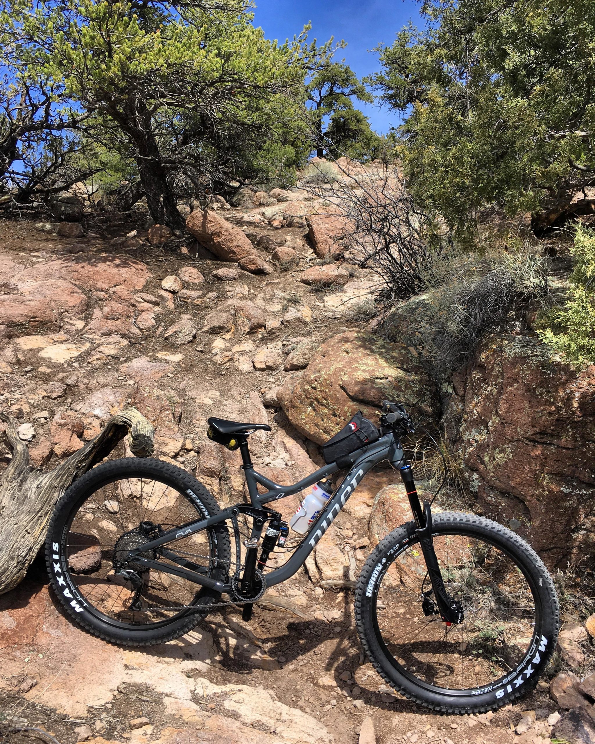 Mountain bike resting on a rocky trail surrounded by greenery and blue skies. The trail features uneven terrain with large stones and sparse underbrush. The bike is black with visible branding on its tire. Unkle Nazty mountain bike trail.