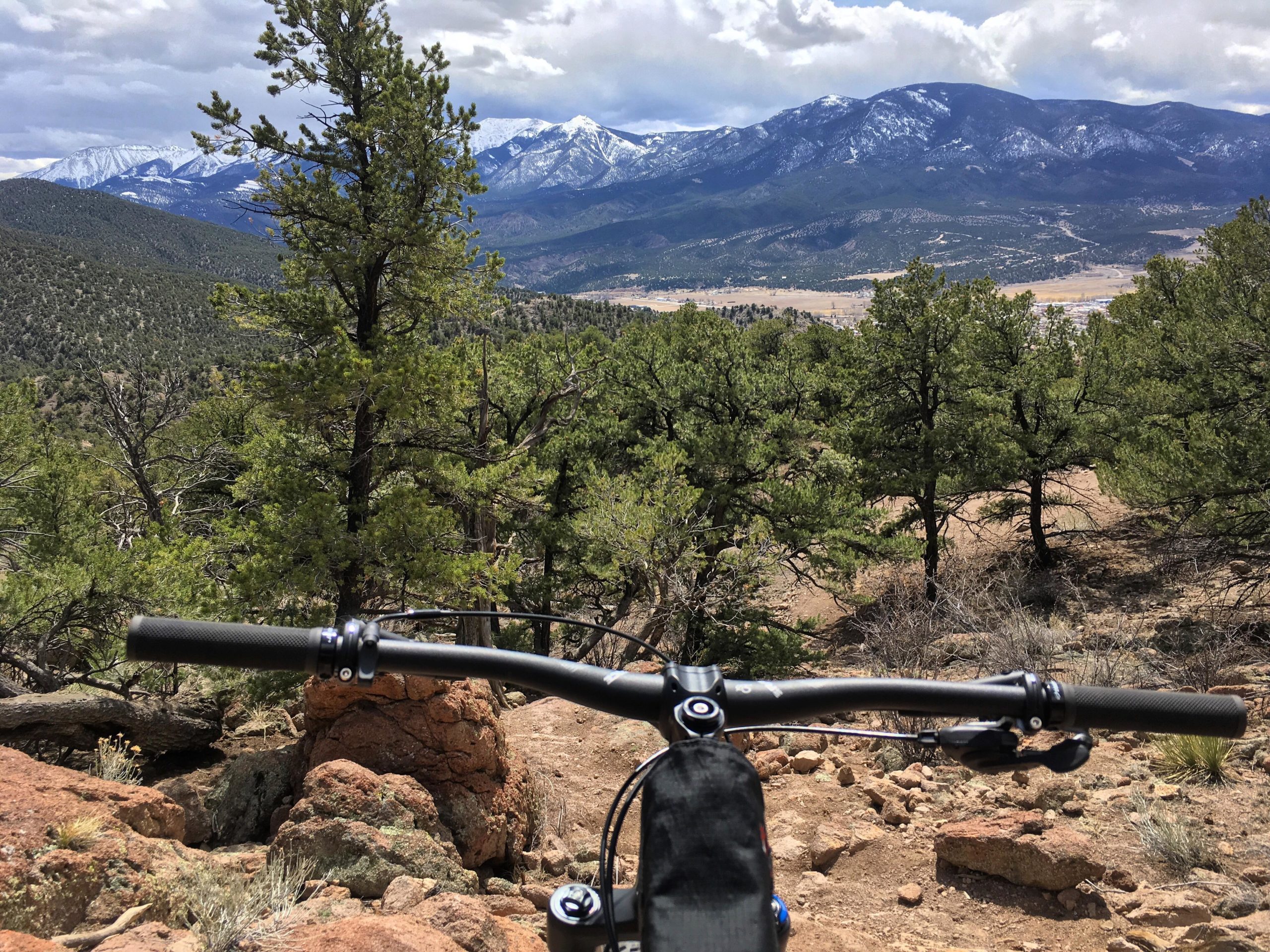 A mountain biker's perspective from the handlebar view, overlooking a rocky terrain with green trees and distant snow-capped mountains under a partly cloudy sky. Unkle Nazty mountain bike trail.