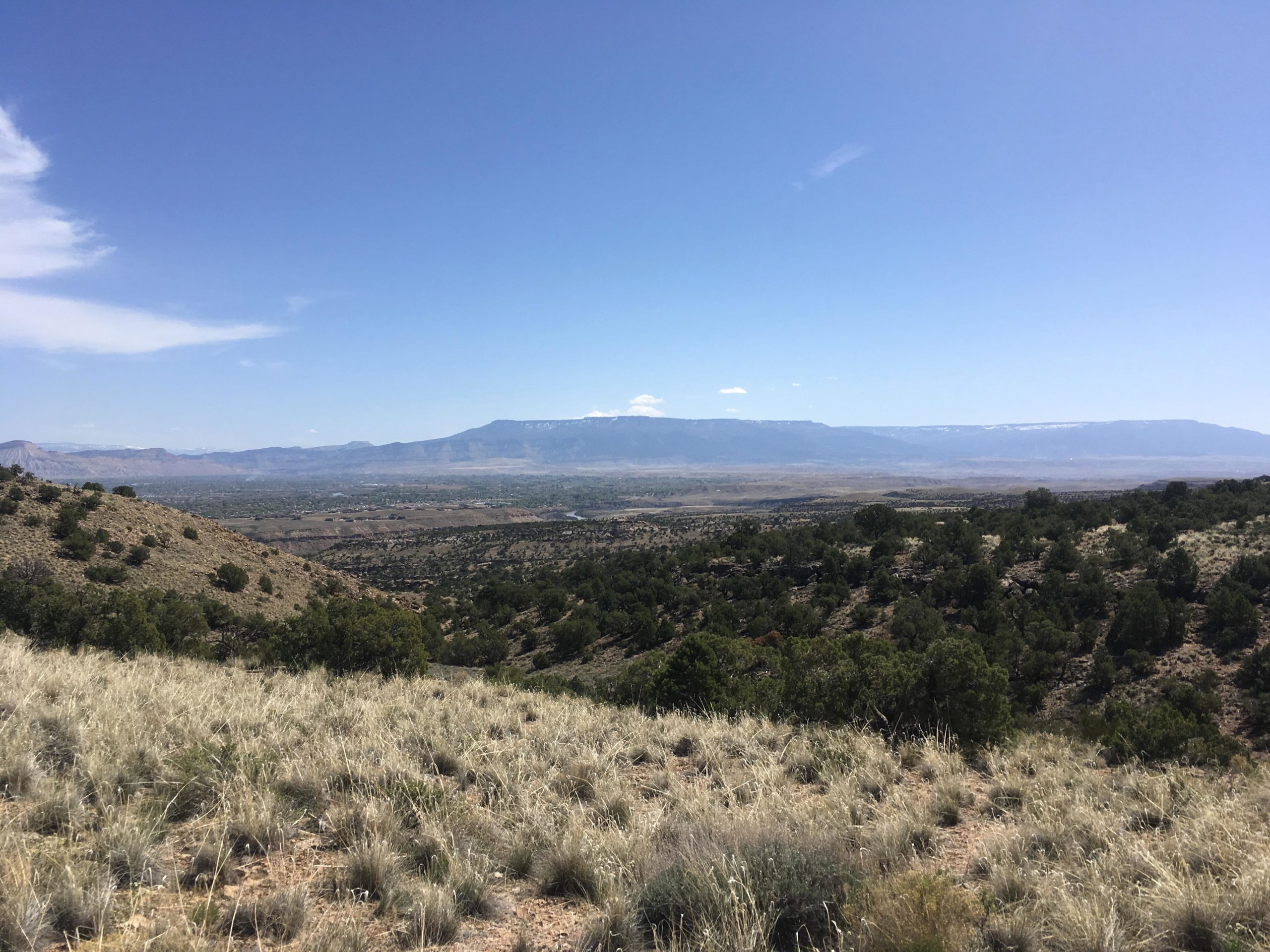 A panoramic view of a vast, arid landscape featuring rolling hills and sparse vegetation under a clear blue sky. In the distance, a mountain range can be seen, with some peaks showing white snow. The foreground consists of dry grass and low shrubs, capturing the essence of a natural outdoor setting. 18 Road Trails / North Fruita Desert mountain bike trail.