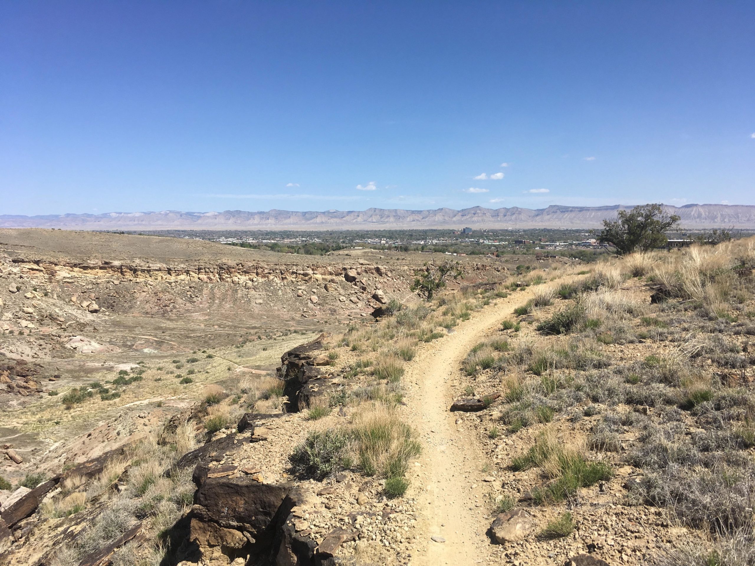 A dirt pathway winds through a hilly landscape, flanked by rocky terrain and sparse vegetation. In the background, mountains stretch across a clear blue sky, while a valley with green trees and buildings is visible below. The scene captures the serenity of nature and the expansive beauty of the outdoors. 18 Road Trails / North Fruita Desert mountain bike trail.