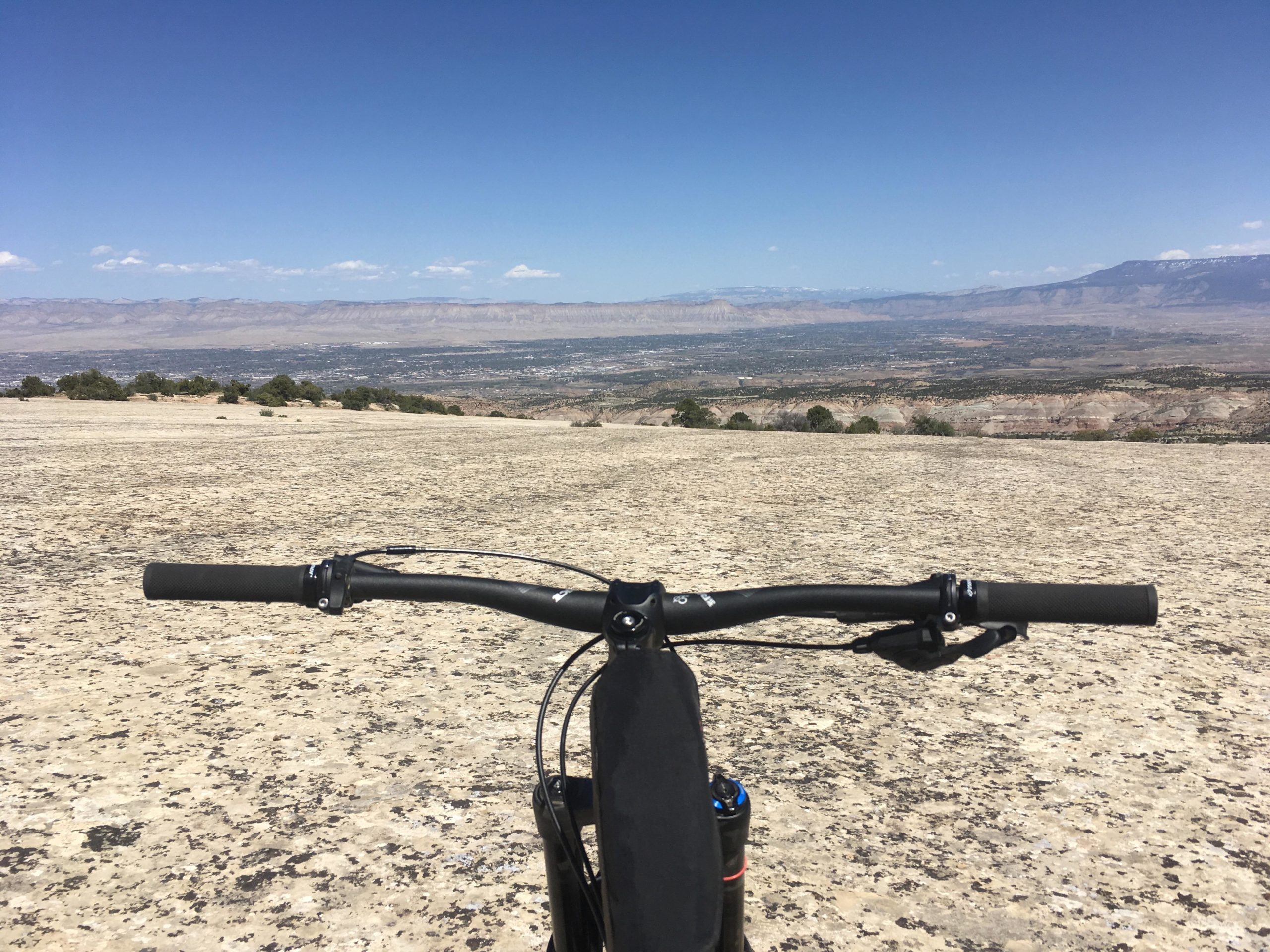 Mountain bike handlebars in the foreground, with a panoramic view of a vast valley and mountains under a clear blue sky in the background. The rocky terrain is visible beneath the bike, emphasizing a sense of adventure and exploration. The Ribbon mountain bike trail.