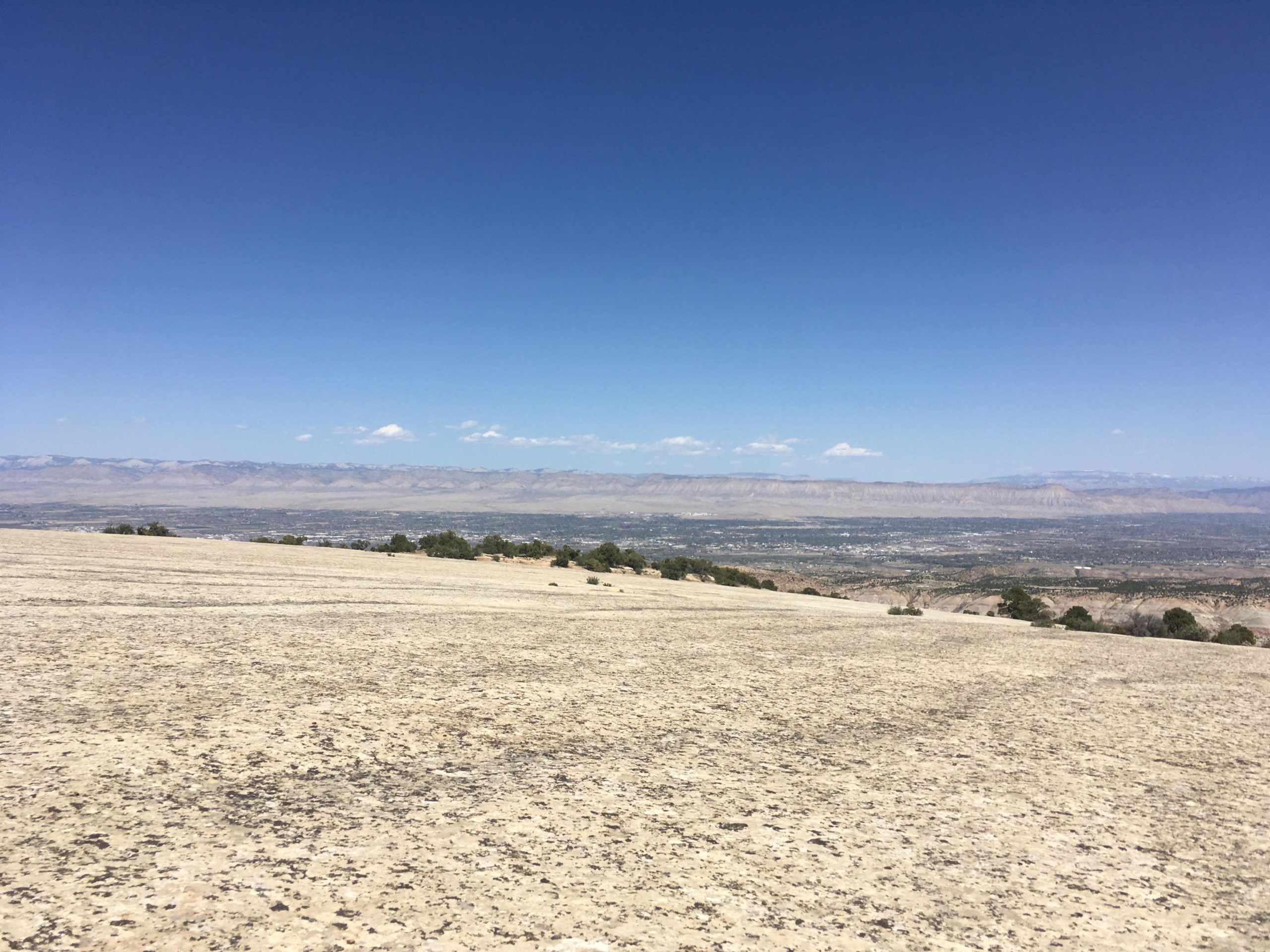 A panoramic view of a rocky landscape under a clear blue sky, with distant mountains and a valley below. The foreground features a smooth, beige rocky surface, and a few scattered trees can be seen on the horizon. The Ribbon mountain bike trail.