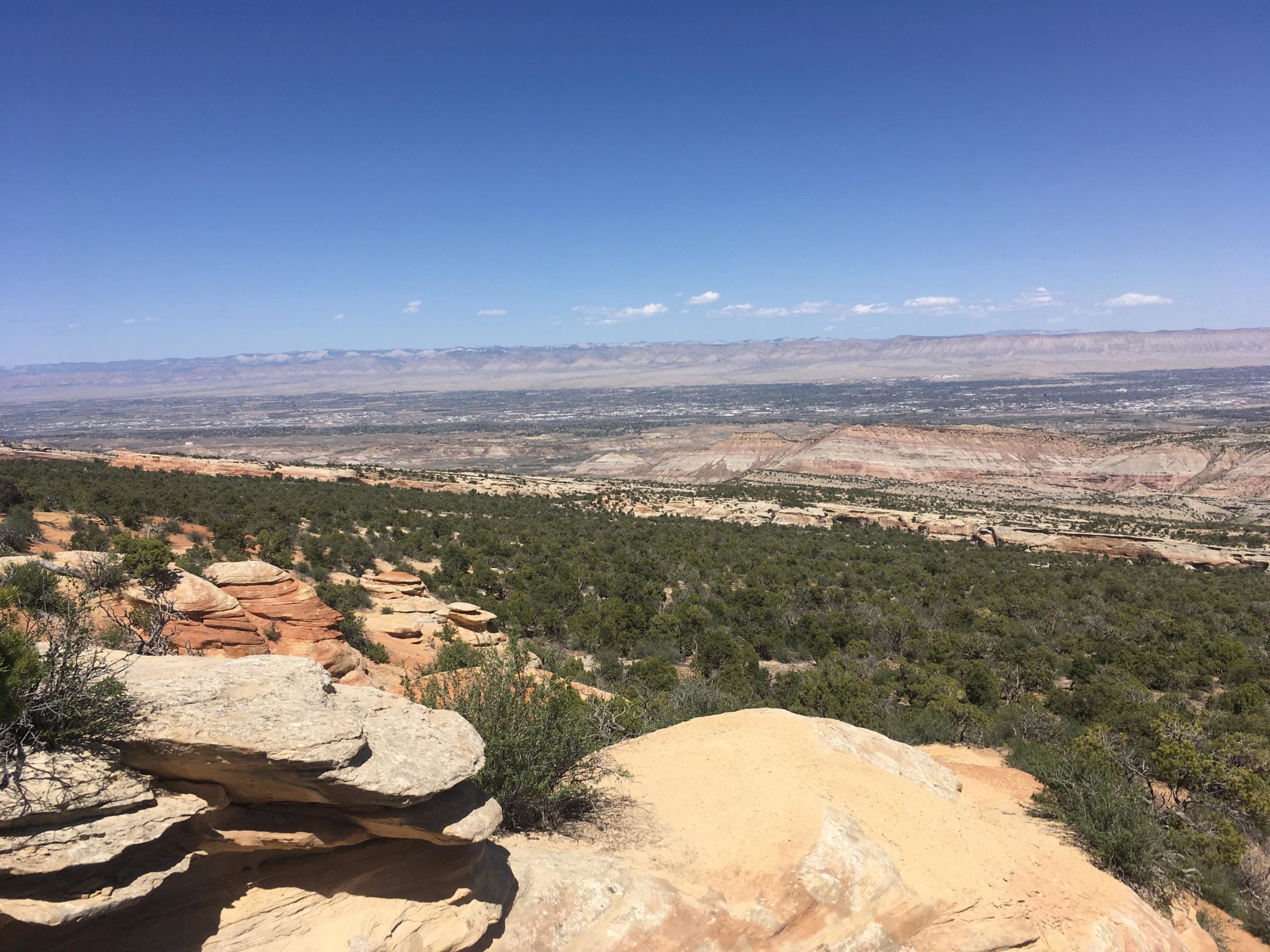 A panoramic view of a mountainous landscape featuring layered rock formations and scattered vegetation, under a clear blue sky. The scene captures rolling hills that gradually descend into the valley, showcasing shades of tan, brown, and green. The Ribbon mountain bike trail.
