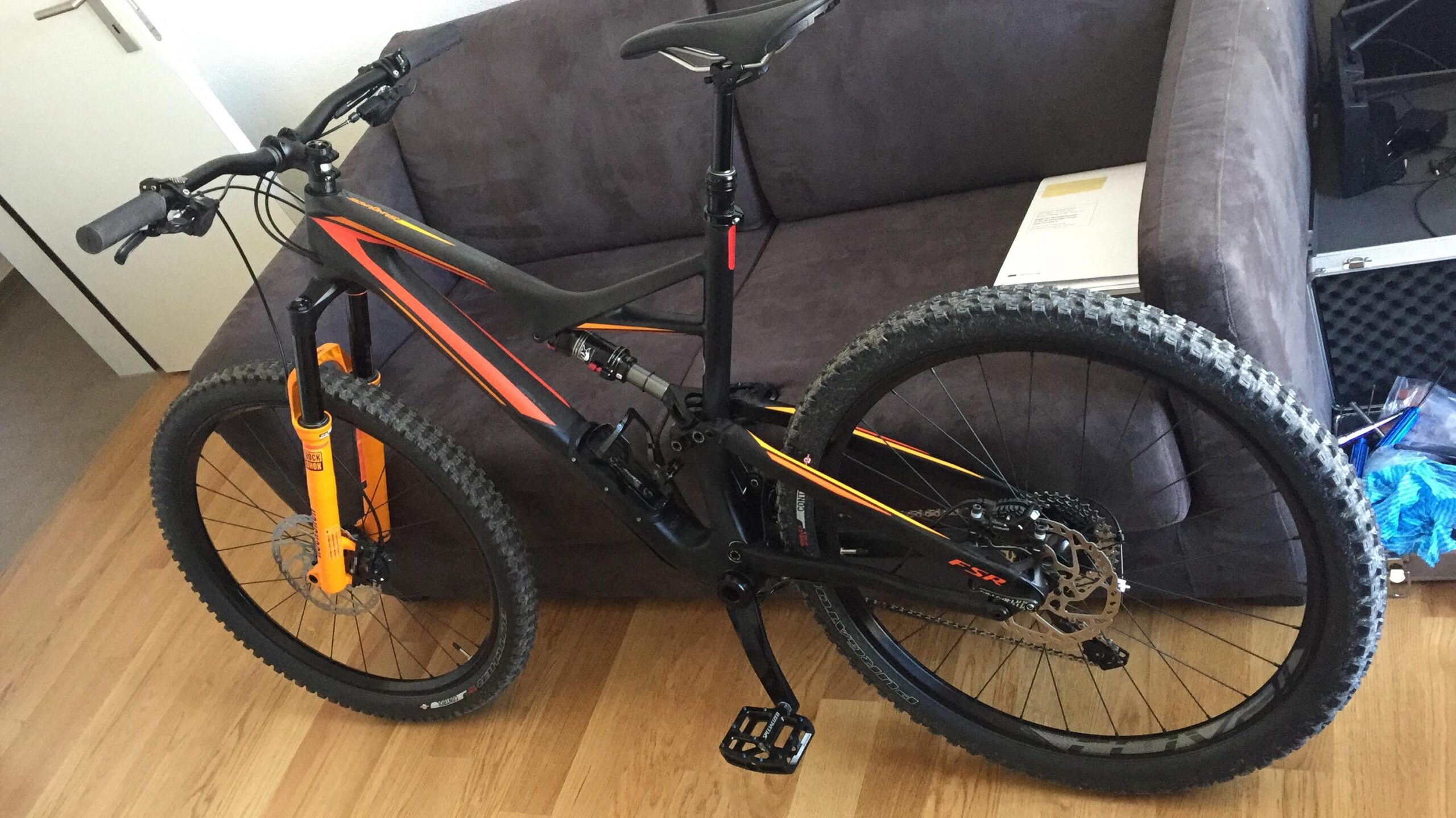 Specialized Stumpjumper FSR comp carbon 650B: A black and orange mountain bike leaning against a brown couch, featuring thick tires and a prominent front suspension fork. The bike's pedals and drivetrain are clearly visible, with a wooden floor and some papers and equipment visible in the background.