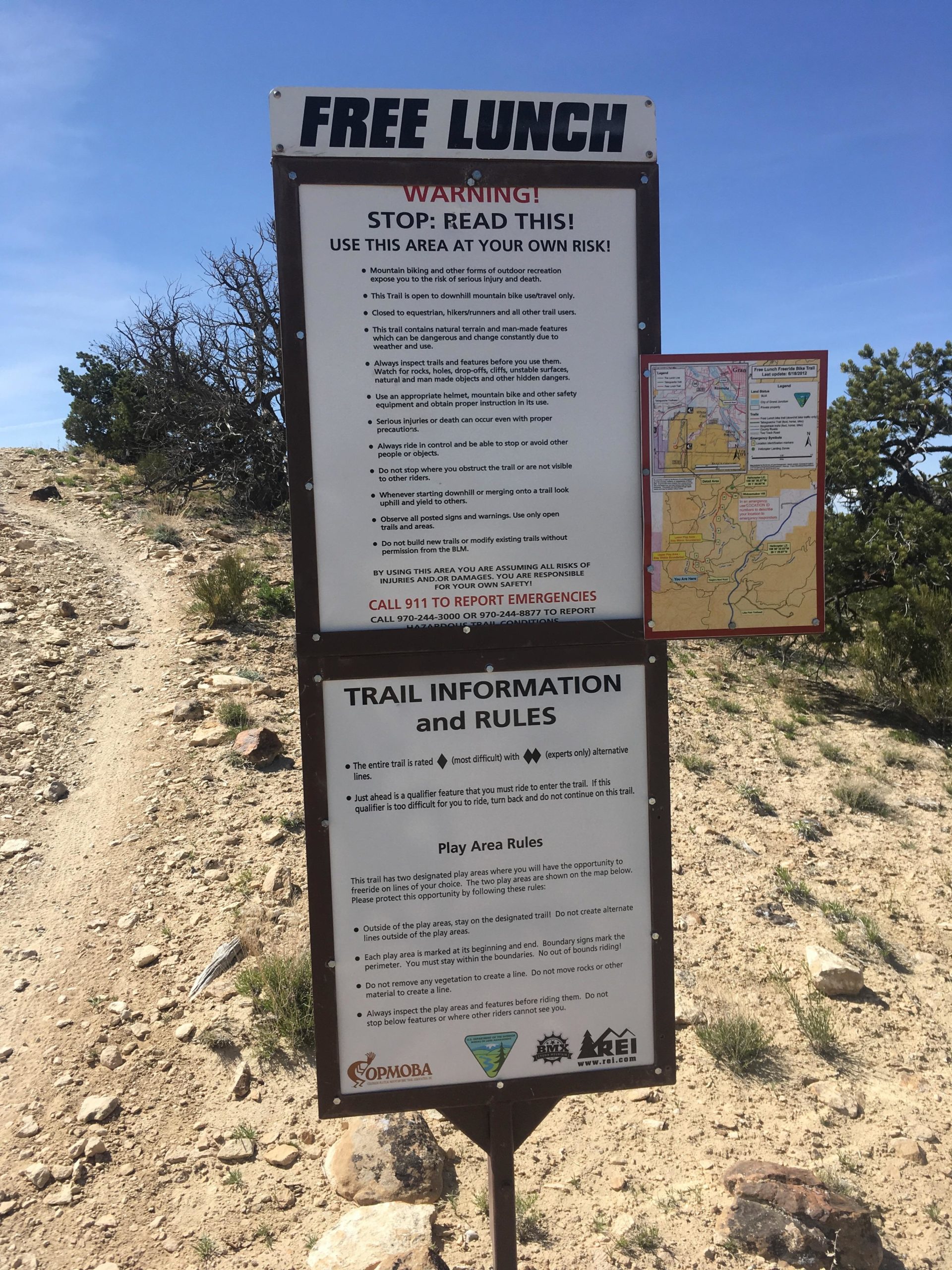 A sign featuring warnings and trail information for a mountain biking area, titled "FREE LUNCH." It includes safety instructions for users, emphasizes the risks involved, and outlines play area rules. The sign is situated on a dirt path surrounded by sparse vegetation and rocky terrain, with a map attached. Clear blue skies are visible above. Free Lunch mountain bike trail.