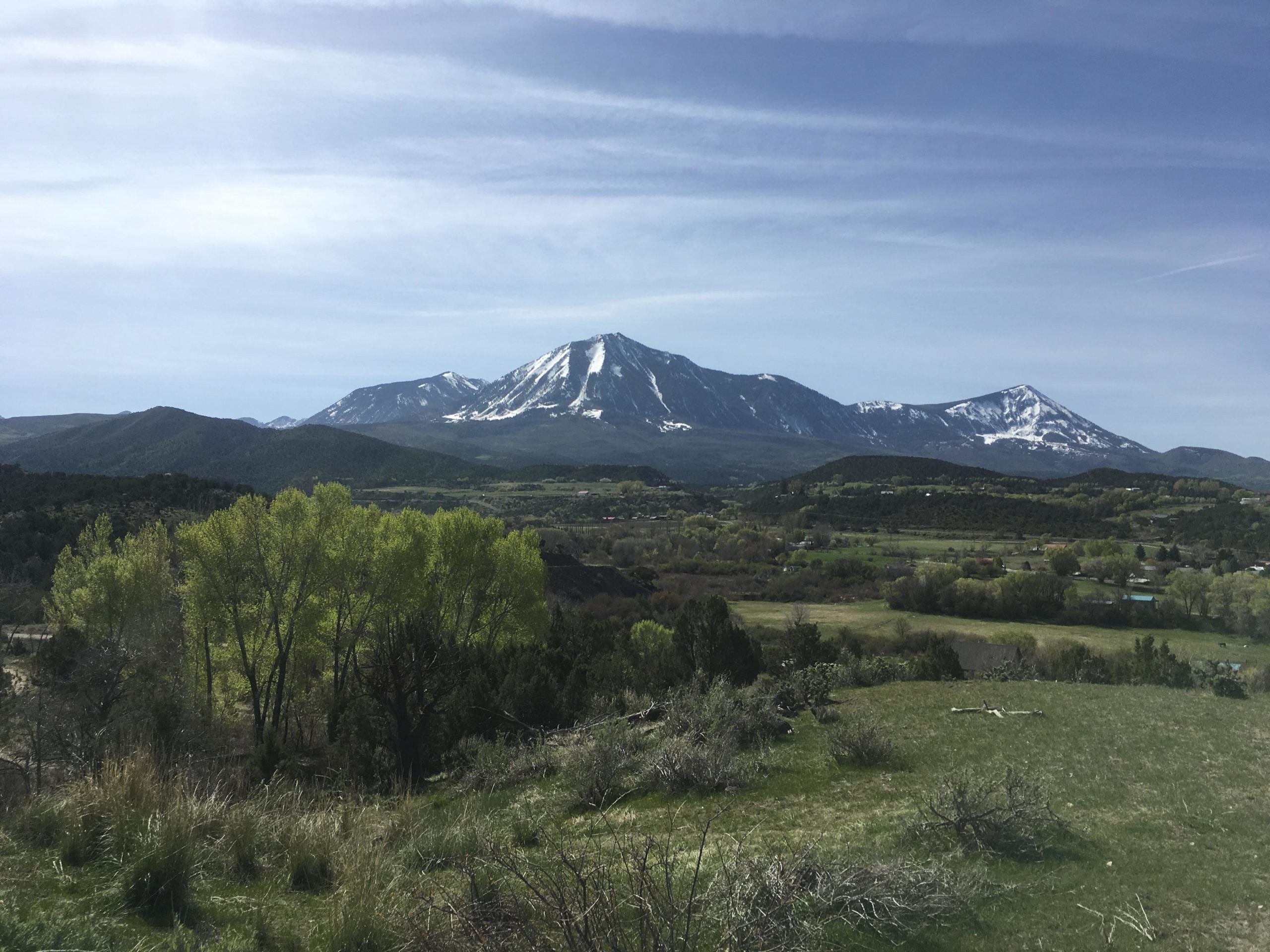 A scenic view of mountains with snow-capped peaks in the background, surrounded by green hills and valleys. The foreground features lush trees and grass, under a clear blue sky with light clouds. Jumbo Mountain mountain bike trail.