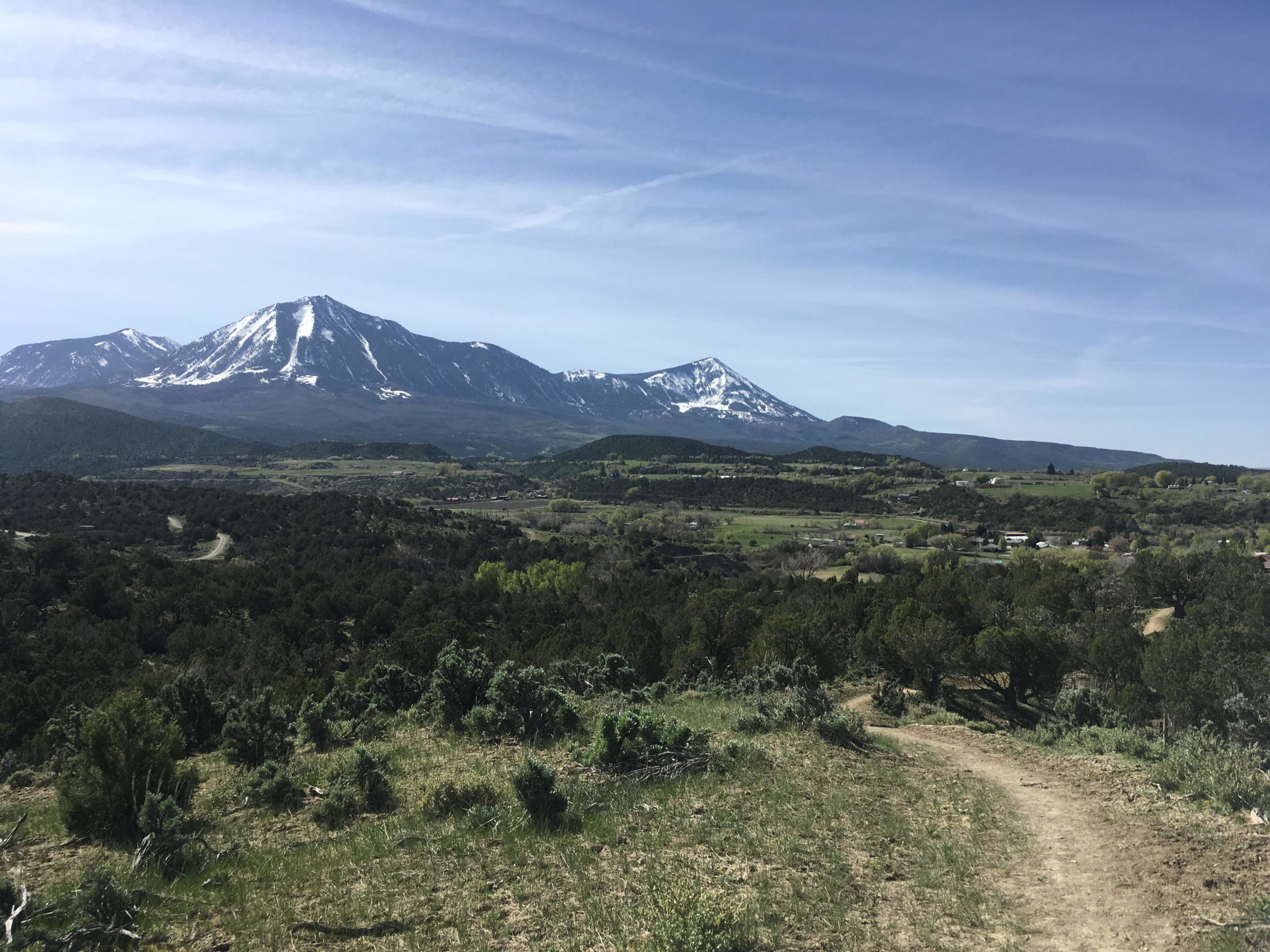A panoramic view of snow-capped mountains overlooking a lush green valley. The landscape features rolling hills dotted with trees and a winding dirt path leading through the foreground. The sky is clear with wispy clouds, suggesting a sunny day. Jumbo Mountain mountain bike trail.