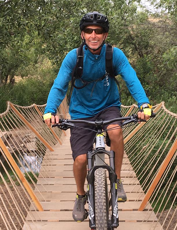 Giant Anthem Advanced 27.5 1: A smiling man in a blue long-sleeve shirt and helmet rides a mountain bike across a wooden bridge surrounded by greenery.