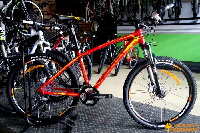 Merida Challenger 800: A vibrant red mountain bike with orange accents, displayed on a stand in a bicycle shop. The bike features thick tires, a sturdy frame, and a front suspension fork. Various other bicycles can be seen in the background, along with a colorful green wall. The floor has a textured surface.