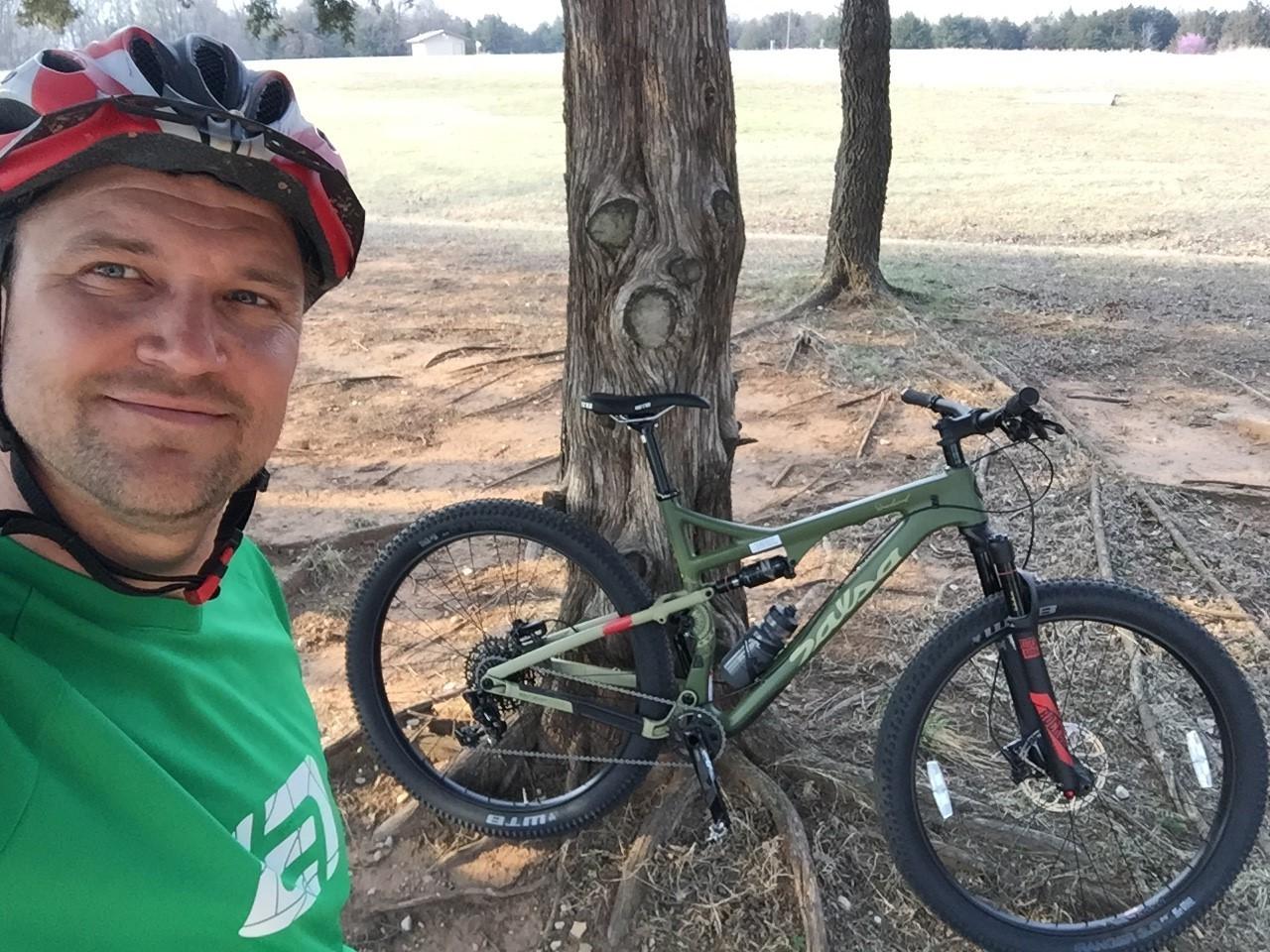 Salsa Deadwood Sus: A person wearing a red and black helmet and a green shirt stands next to a mountain bike leaning against a tree in a grassy area. The background features a field and trees.