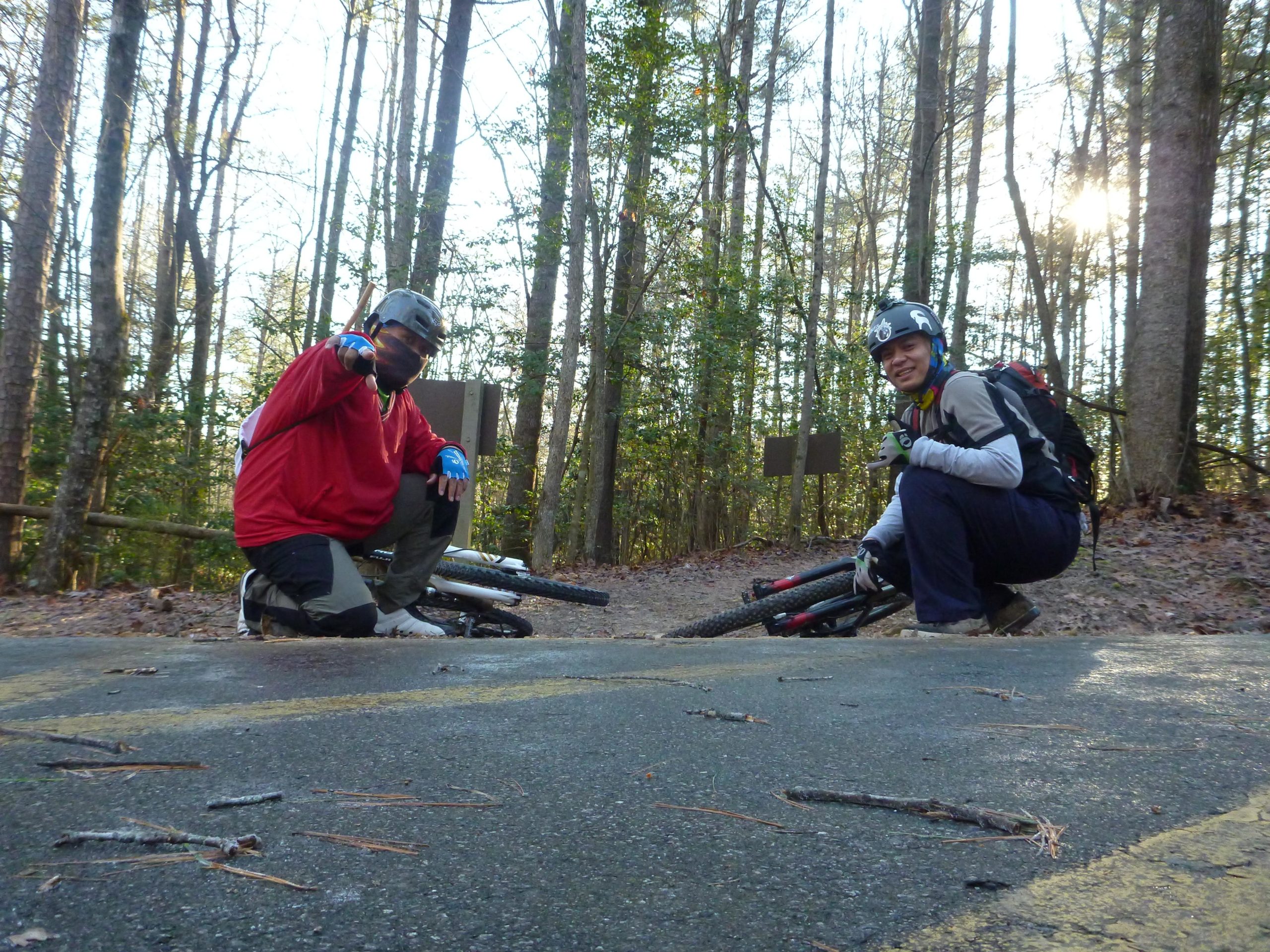 Two mountain bikers posing on a forest trail, one wearing a red jersey and helmet, the other in gray attire, both crouching beside their bikes. The background features tall trees and a hint of sunlight breaking through the leaves. Overmountain Victory Trail mountain bike trail.