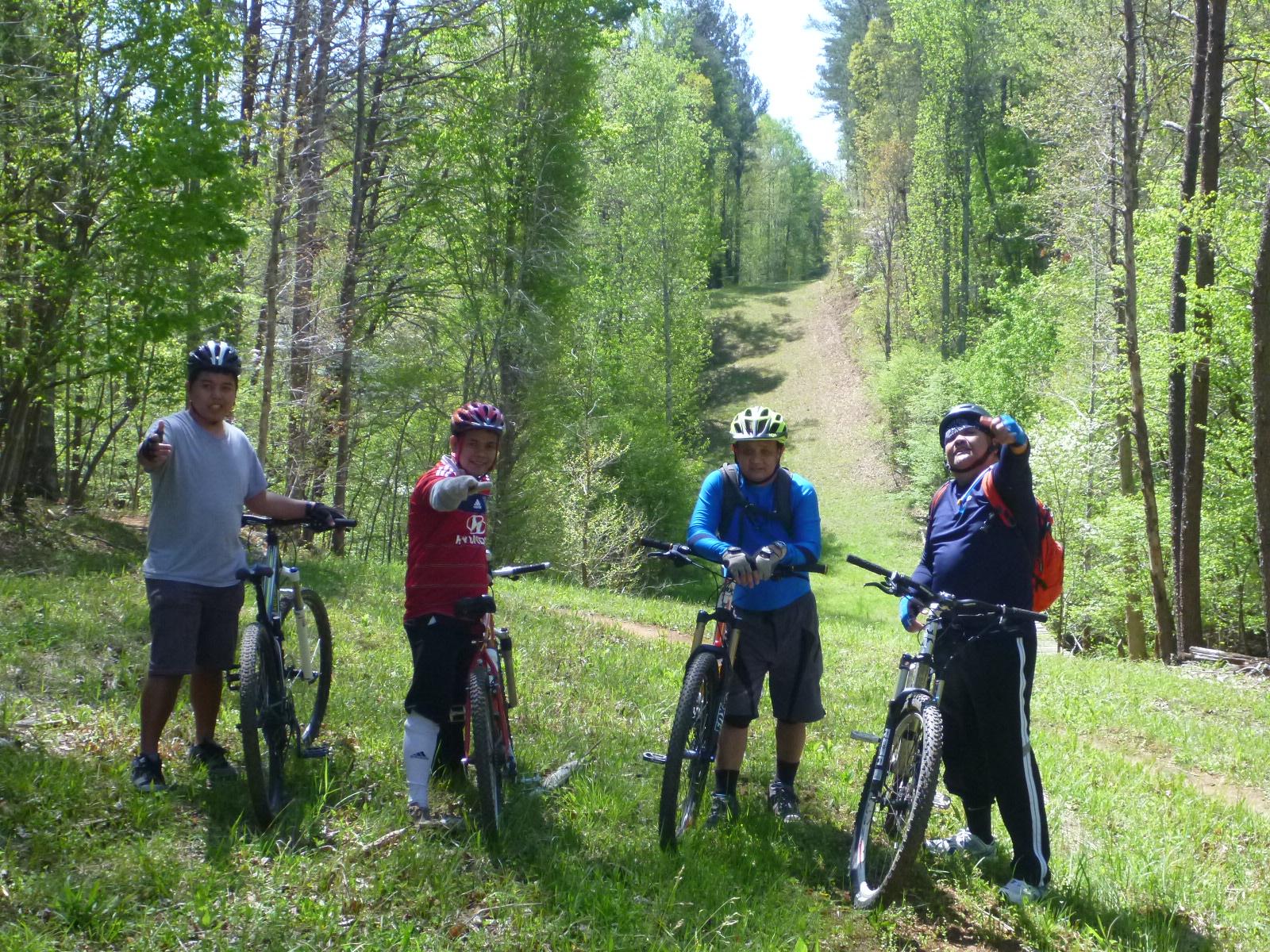 Four people on mountain bikes pose for the camera in a lush, green forest setting. They are wearing helmets and casual biking attire, standing on a grassy path that slopes gently uphill. The group appears to be enjoying their time outdoors, with smiles on their faces while giving a thumbs-up gesture. Surrounding them are tall trees and vibrant foliage, indicating a sunny day. Cedar Ridge Chatmoss mountain bike trail.