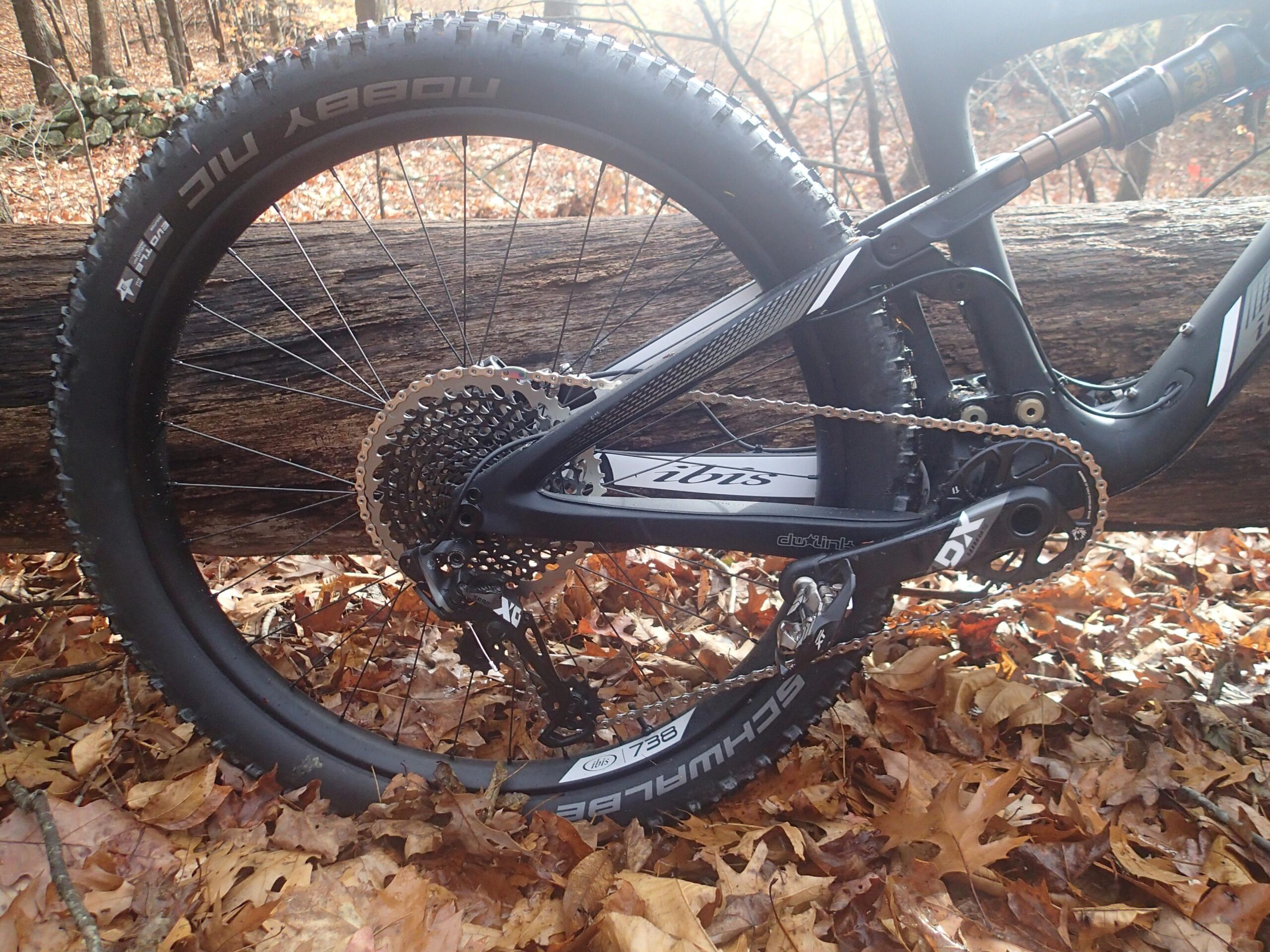 Ibis Mojo 3: Close-up view of a mountain bike's rear wheel and drivetrain, resting on a log covered with autumn leaves. The bike features a black frame with a sleek design, accessible gears, and a high-performance tire. The background includes blurred trees and fallen leaves, suggesting an outdoor setting.