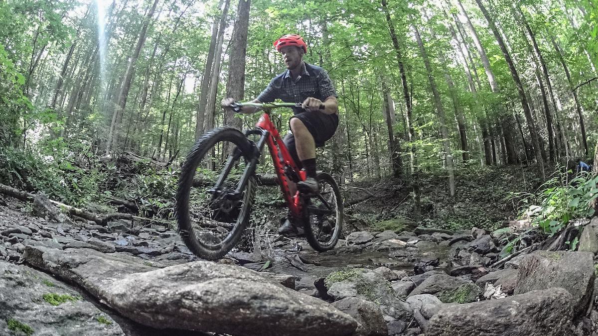 A mountain biker in a red helmet rides over rocky terrain in a lush green forest. Sunlight filters through the trees, illuminating the scene as the cyclist skillfully navigates the natural path. Squirrel Gap mountain bike trail.