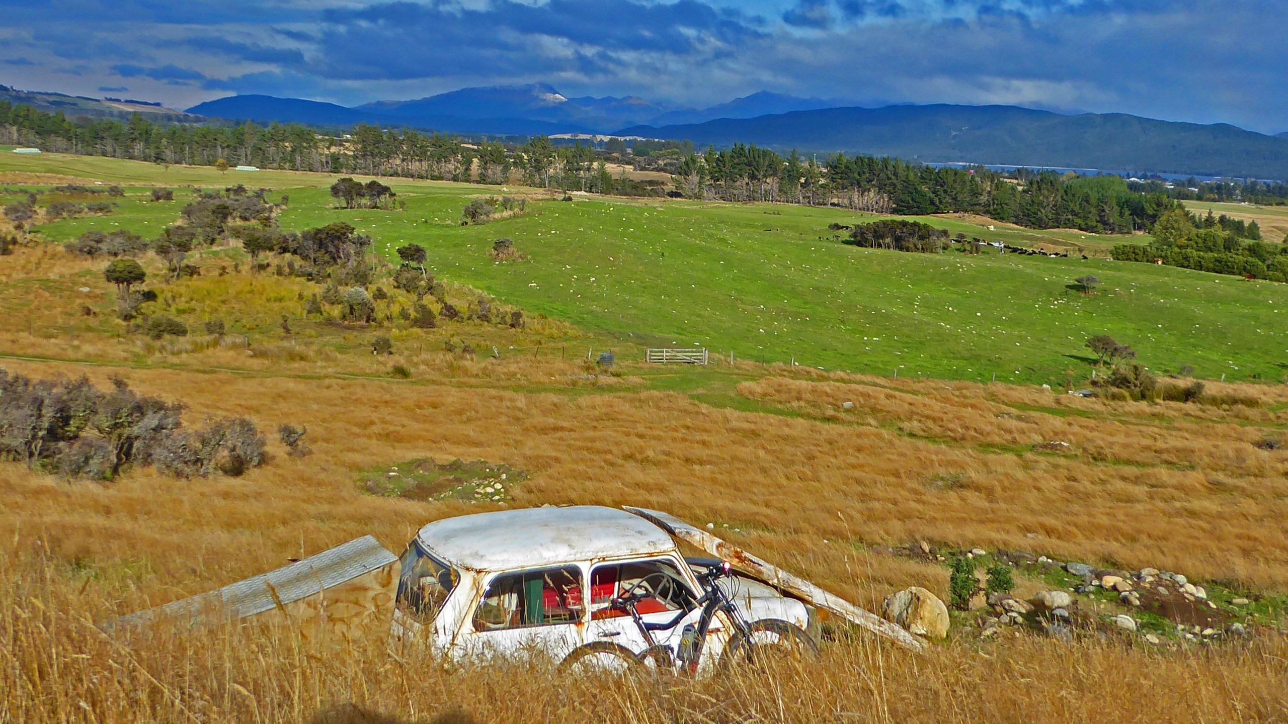 A rural landscape featuring a dilapidated white car with a bicycle leaning against it, set against rolling hills of golden grass and green pastures. In the background, distant mountains are visible under a partly cloudy sky. Perenuka mountain bike trail.