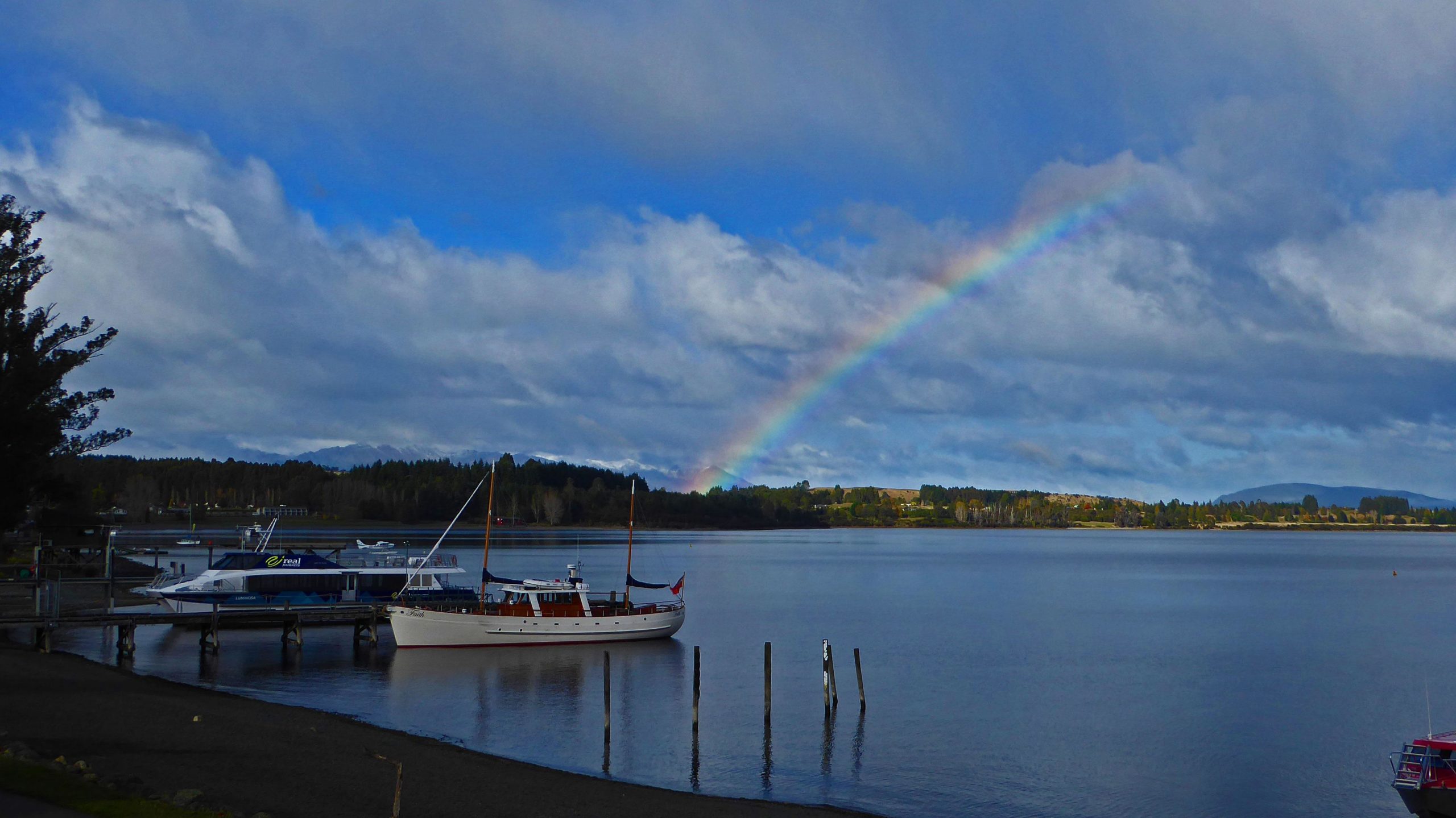 A scenic view of a calm lake featuring several boats docked at a wooden pier. In the background, a vibrant rainbow arches over the green landscape and forested hills under a partly cloudy sky. Te Anau - Manapouri mountain bike trail.