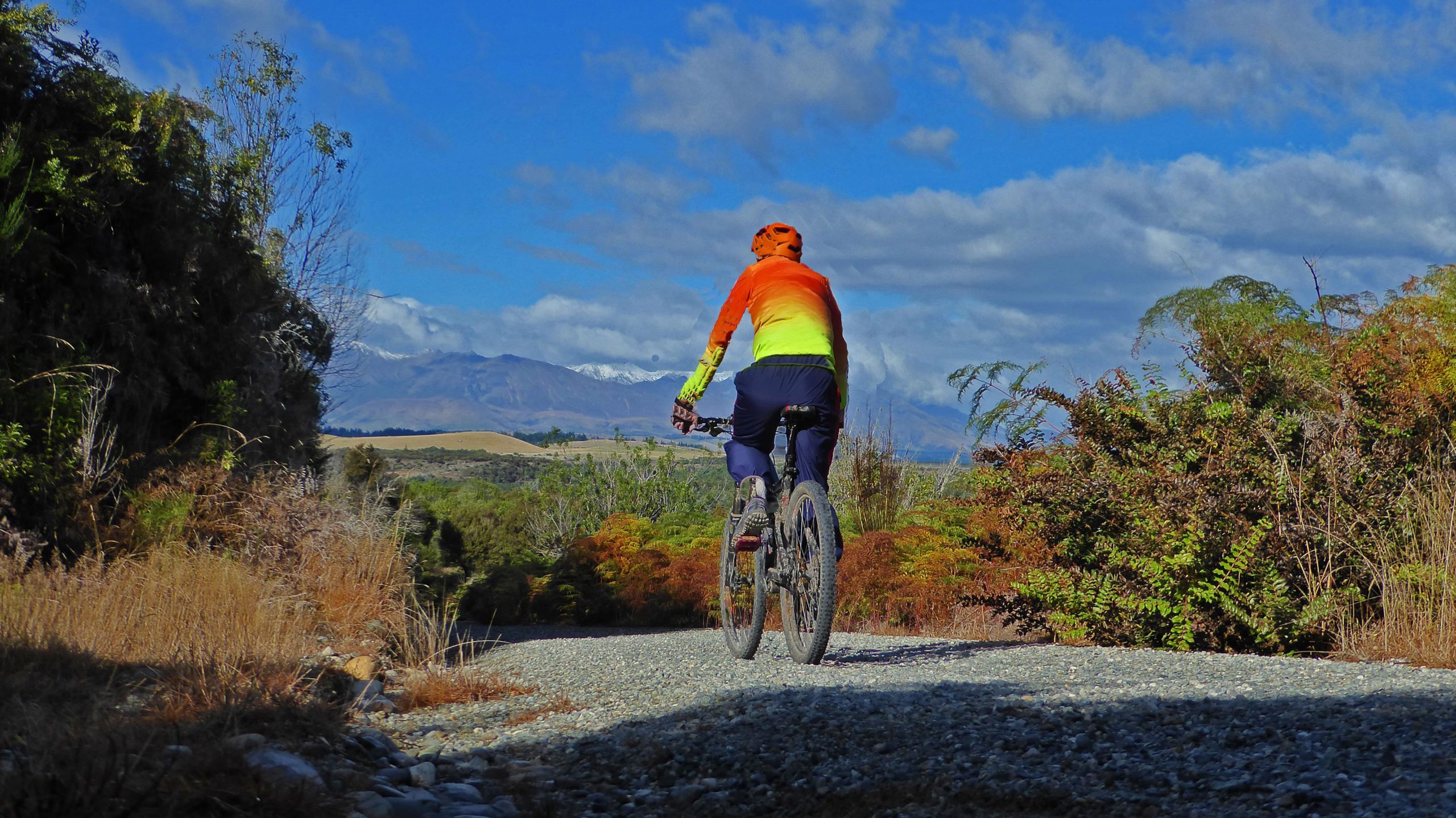 A cyclist in a bright orange and yellow jacket rides along a gravel path surrounded by greenery and bushes, with mountains in the background under a blue sky with scattered clouds. Te Anau - Manapouri mountain bike trail.