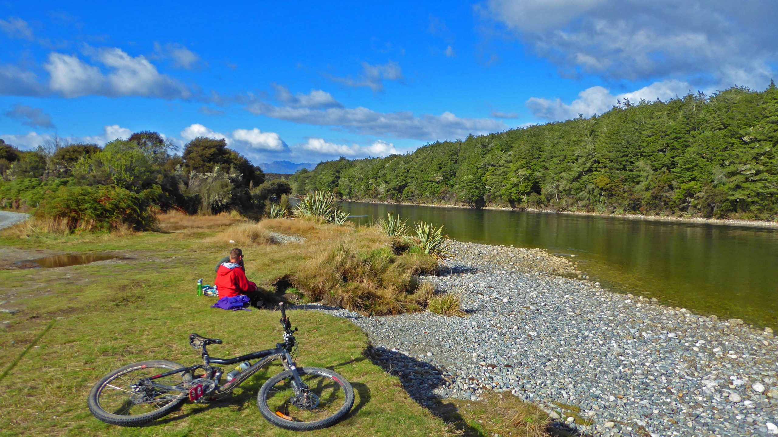 A peaceful riverside scene featuring a person sitting on the grass near the water, surrounded by greenery and trees. A black mountain bike is positioned in the foreground, and the sky is bright with clouds. The tranquil river reflects the surrounding landscape, creating a serene atmosphere. Te Anau - Manapouri mountain bike trail.