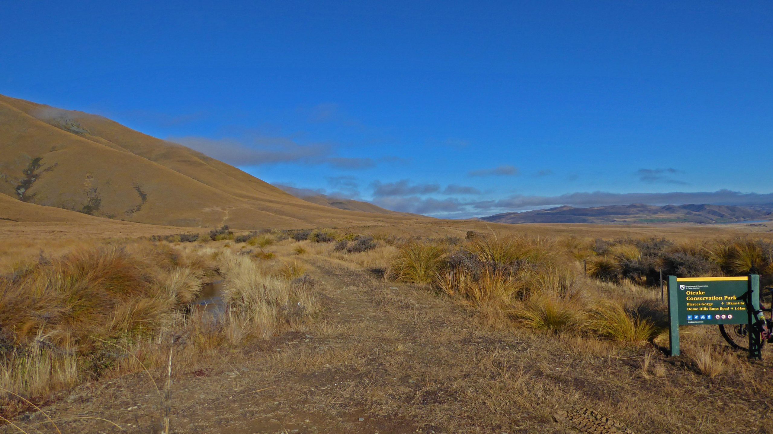 A scenic view of a trail leading through dry grassland, flanked by rolling brown hills under a clear blue sky. A green sign indicates the entrance to Oteake Conservation Park, providing information about nearby trails. Mount Ida Water Race mountain bike trail.