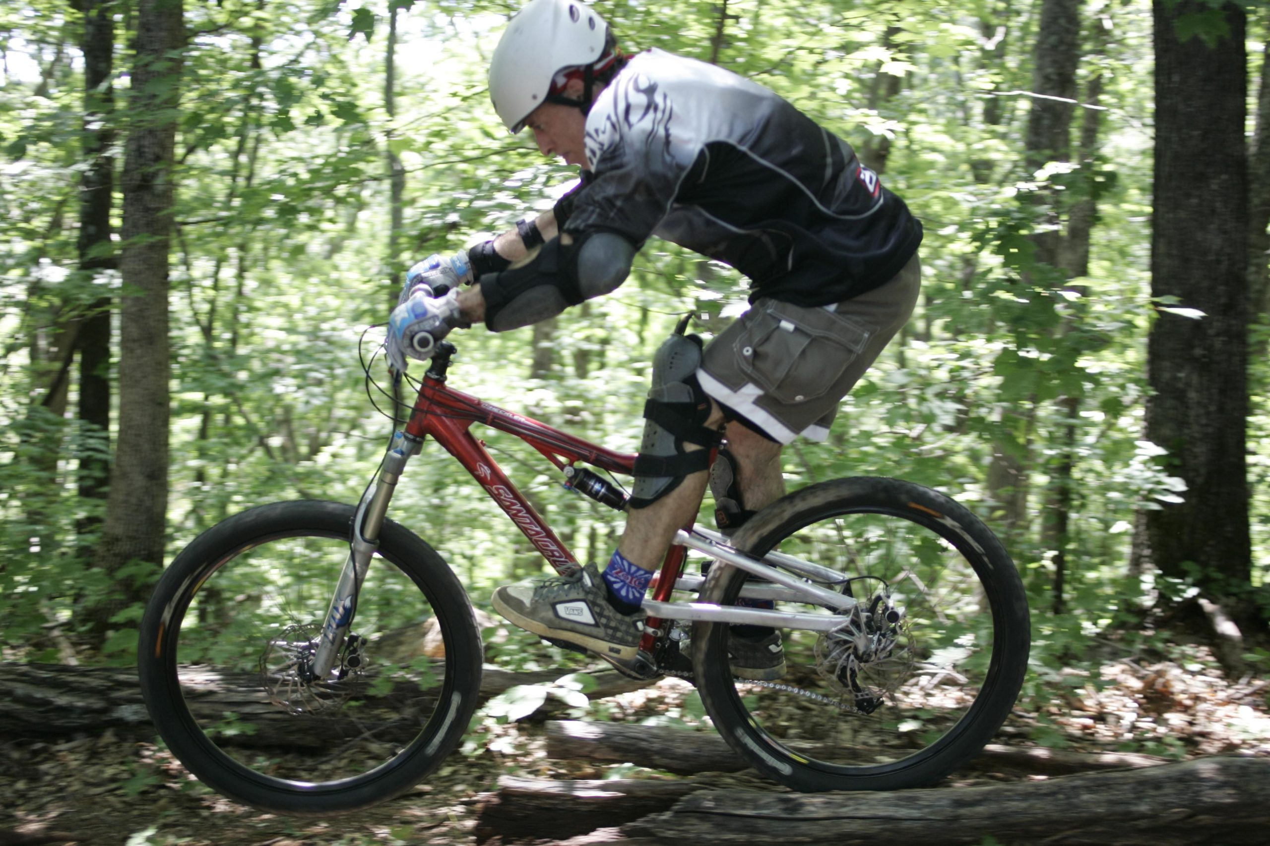 A mountain biker in protective gear rides over a log on a forest trail, surrounded by lush greenery. The cyclist is in mid-air, displaying skill and balance on a red mountain bike. Kitsuma mountain bike trail.