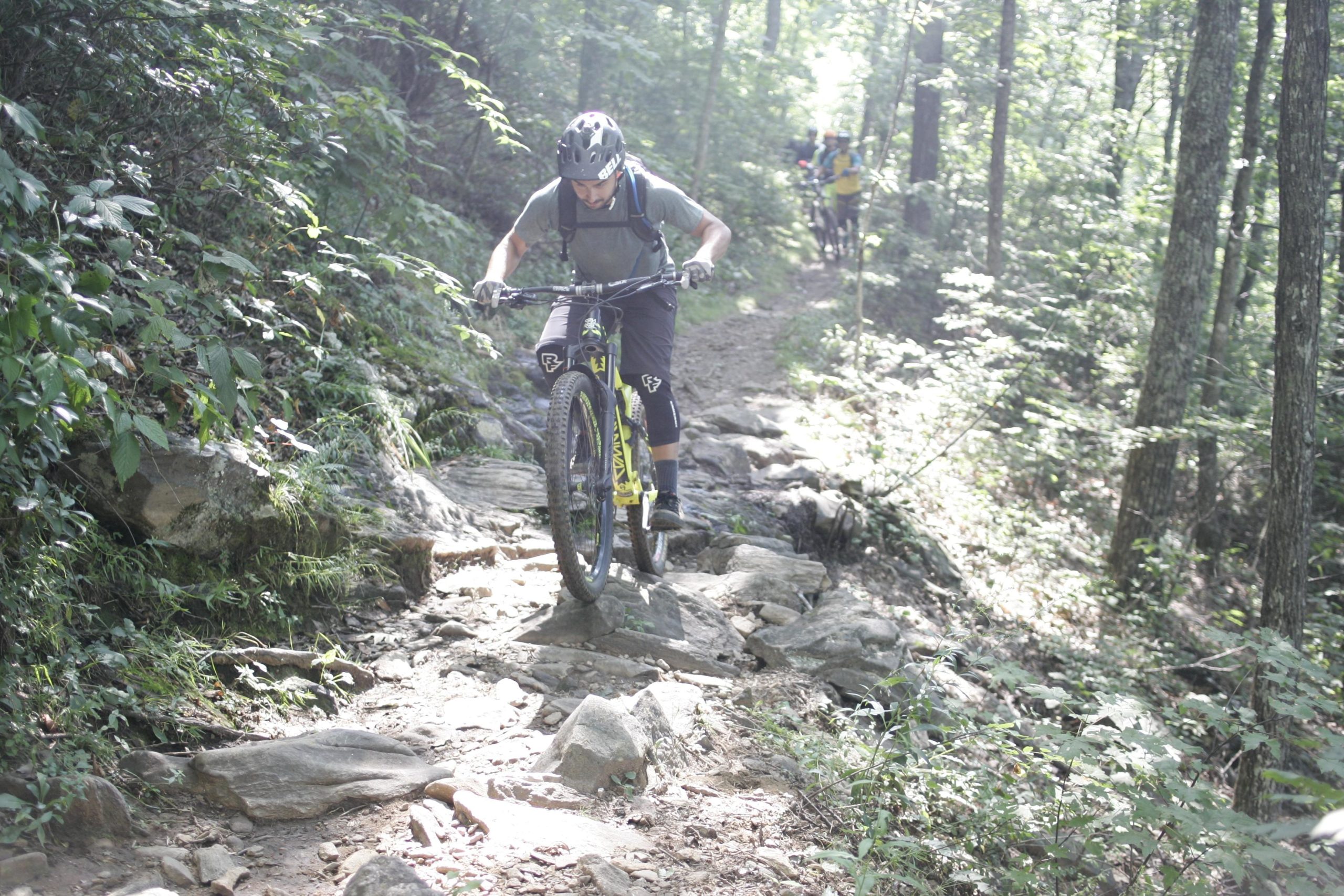A mountain biker maneuvering over rocky terrain in a forested area, with sunlight filtering through the trees. Another cyclist is visible in the background, navigating the trail. Laurel Mountain mountain bike trail.