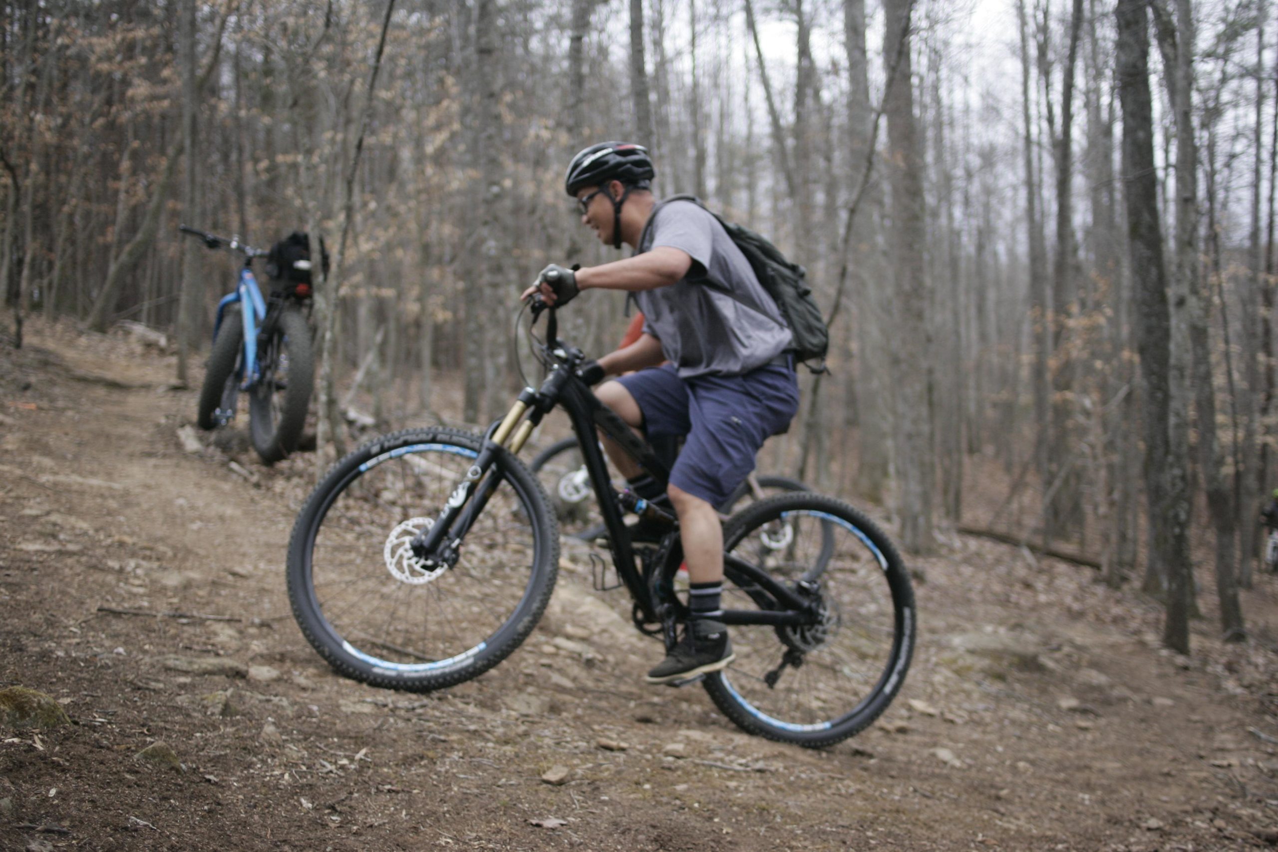 A person riding a mountain bike on a dirt trail in a forested area, performing a wheelie while wearing a helmet and backpack. Another bicycle is leaning against a tree in the background. The scene is set in a wooded environment with sparse foliage. Mountain Laurel Trails mountain bike trail.