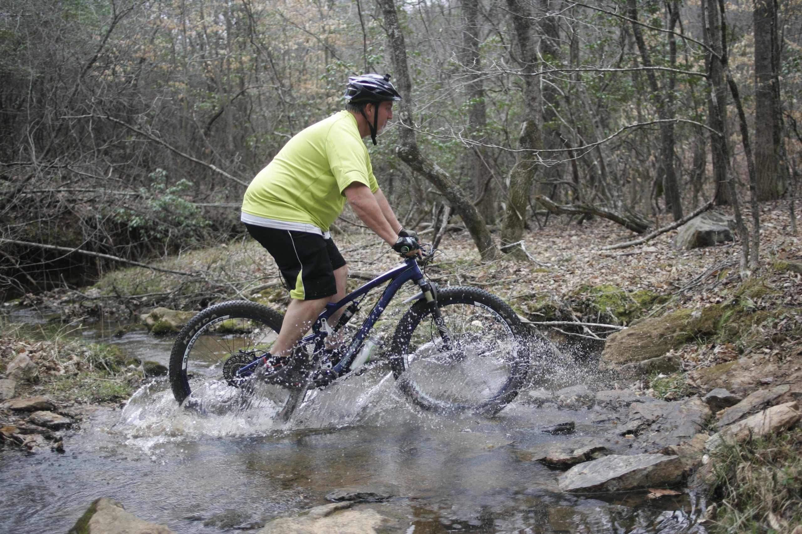 A cyclist in a bright yellow shirt rides through a shallow stream on a mountain bike, splashing water as they navigate over rocky terrain surrounded by trees and foliage. Mountain Laurel Trails mountain bike trail.